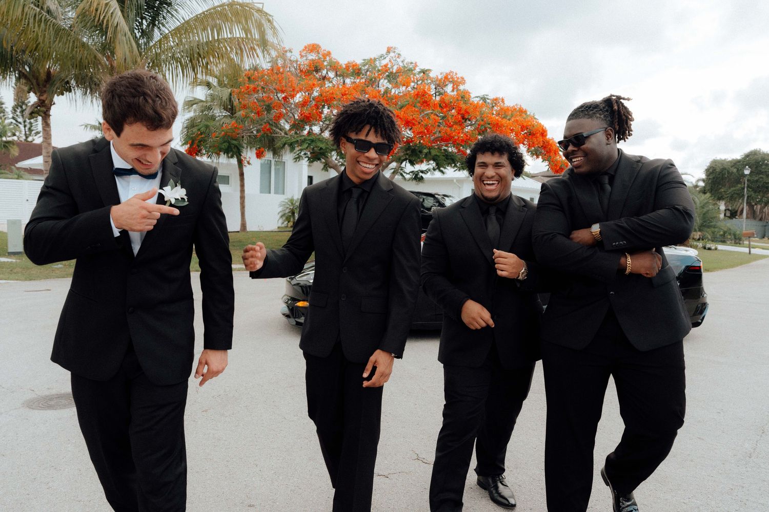 Four men in black suits laughing and posing outdoors near a flowering tree at a formal event.