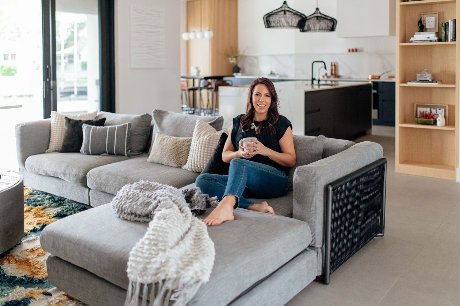 Woman relaxing on a gray sectional sofa in a modern open-plan living room, holding a coffee mug.
