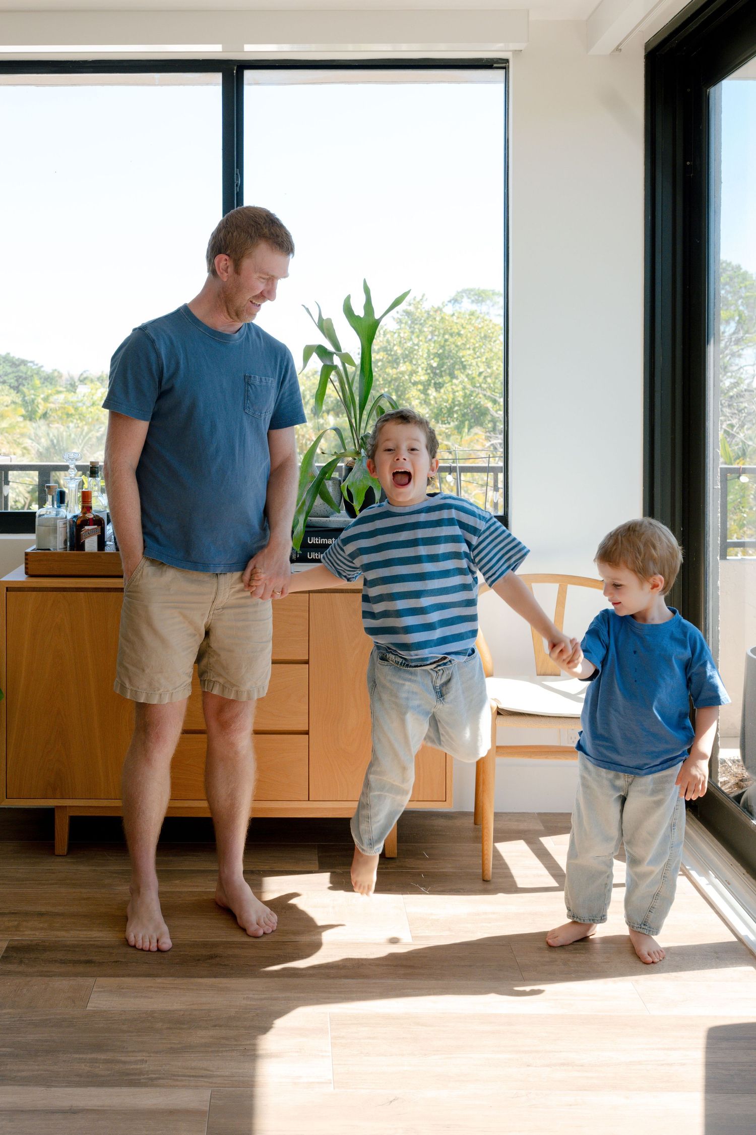 Father and two young boys playing and laughing together in a modern sunlit living room with hardwood floors.