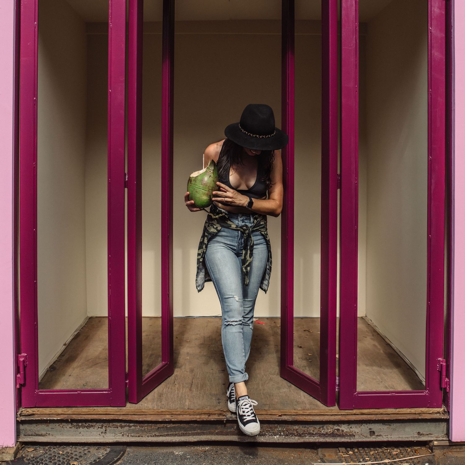 Woman in black hat drinking from a coconut in a pink doorway wearing jeans and sneakers.
