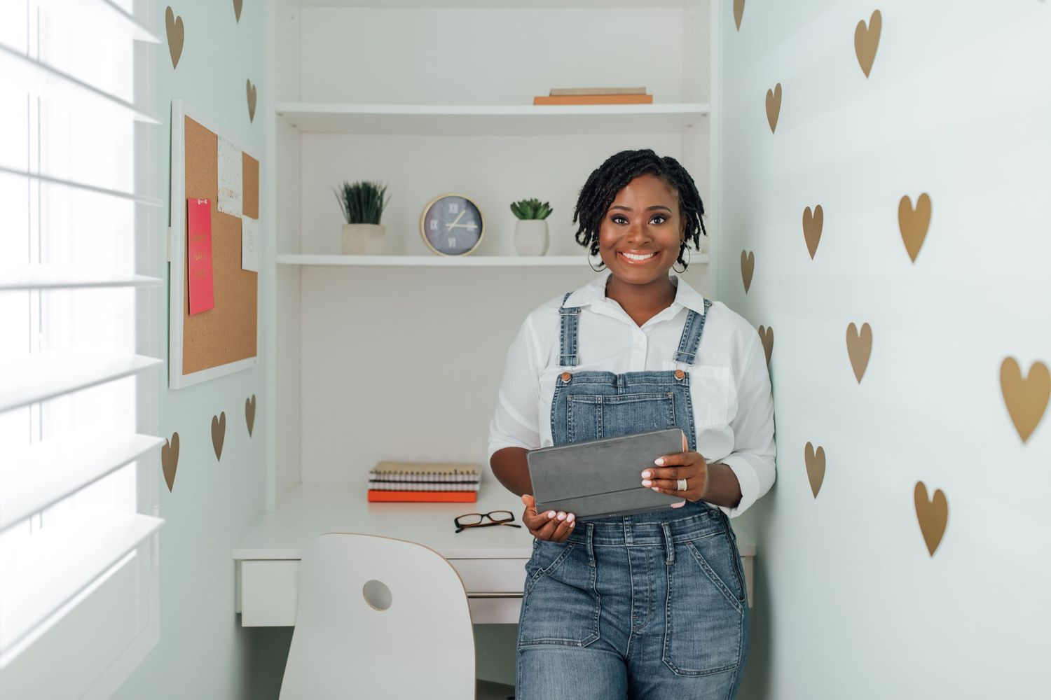 Smiling woman in denim overalls holding tablet in a decorated home office with heart wall decals.