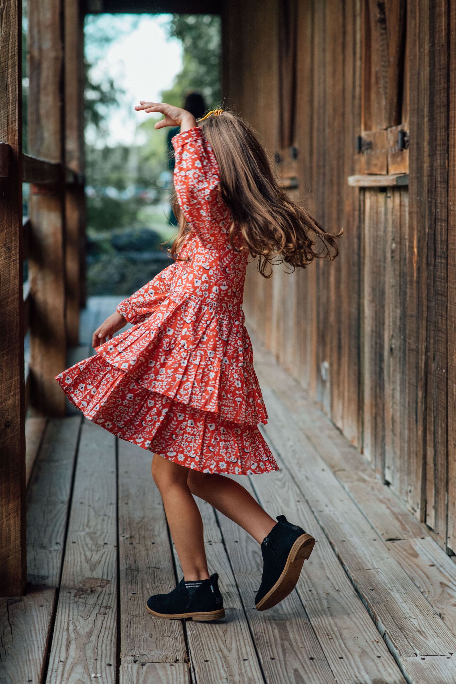 A young girl in a red floral dress and black boots twirling on a rustic wooden porch.