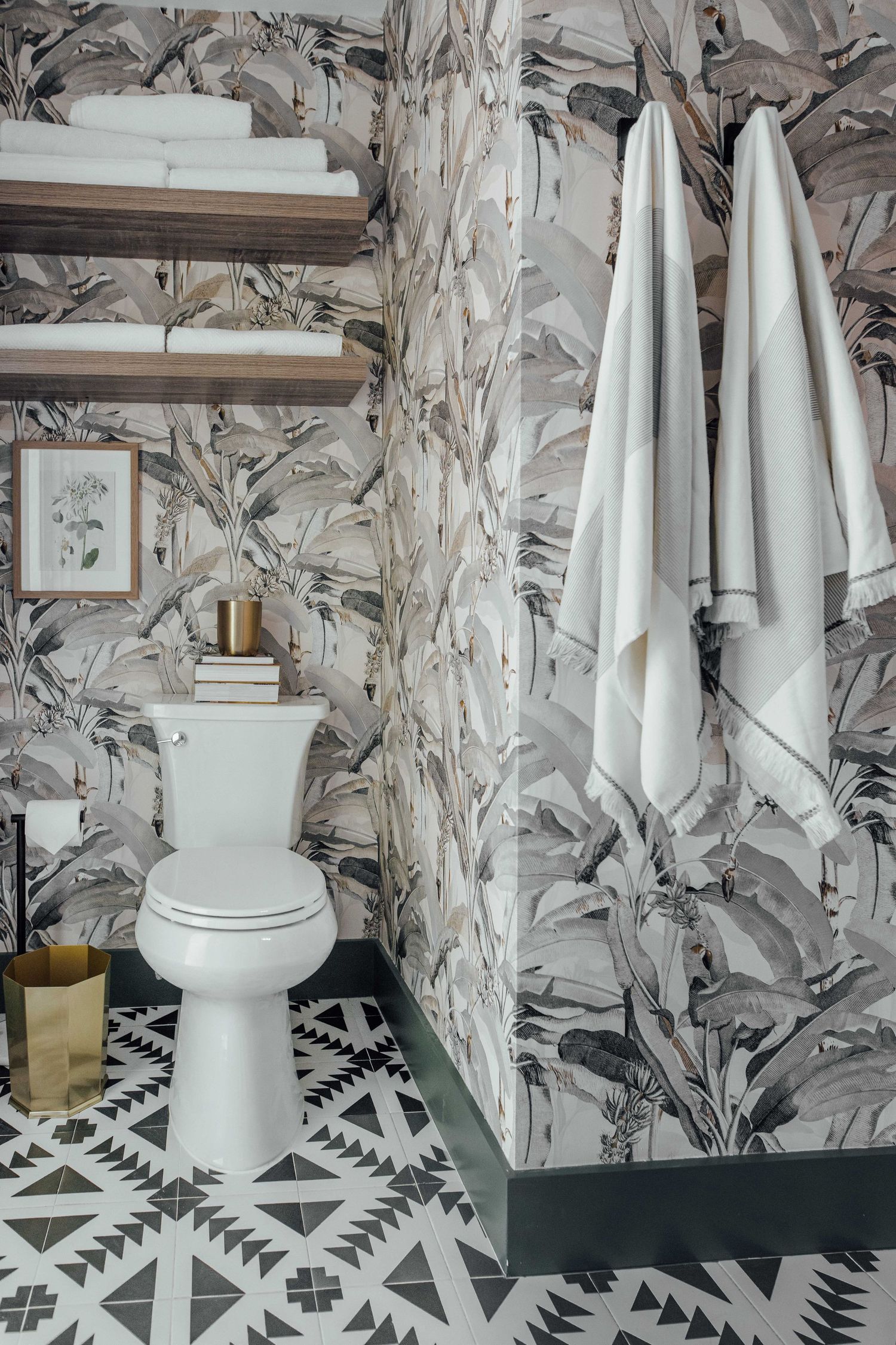 Bathroom with tropical leaf wallpaper, white toilet, wooden shelves, geometric floor tiles, and hanging white towels.