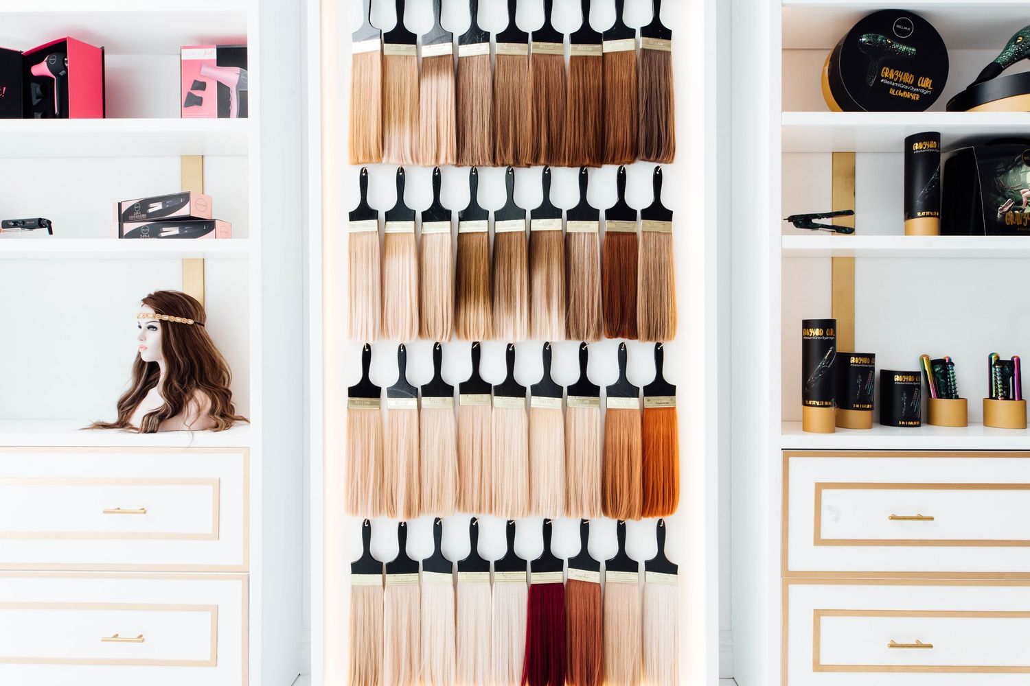 A salon display wall featuring rows of hair extension color swatches in shades from blonde to brunette, flanked by white shelving units.