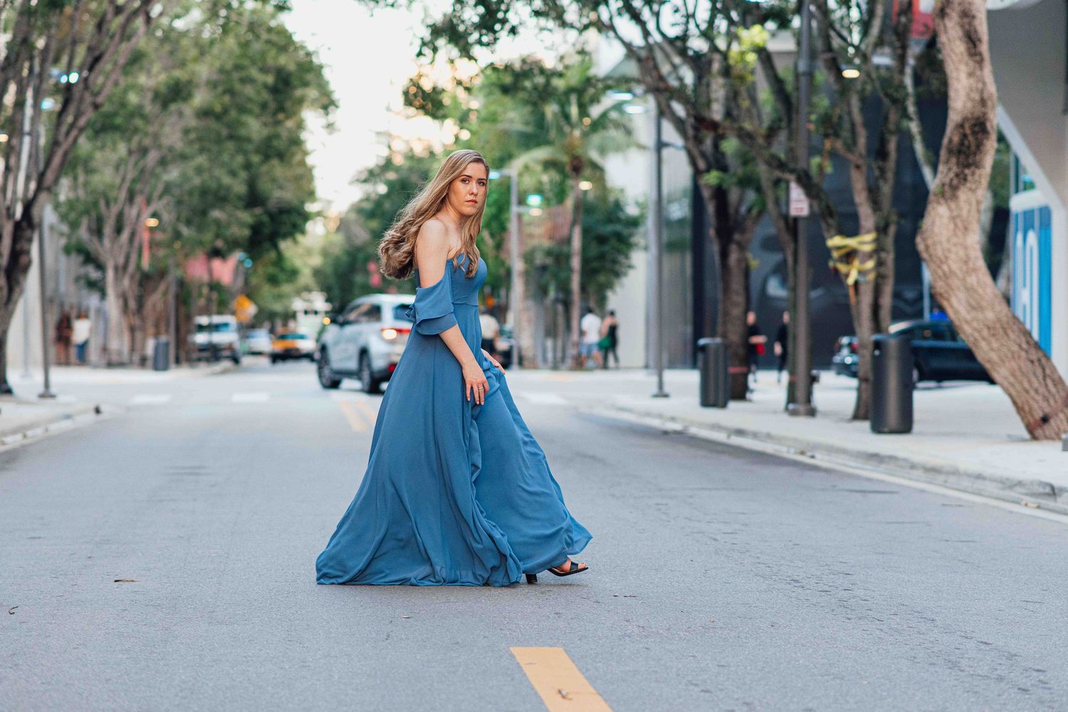 Elegant woman in a flowing blue maxi dress walking gracefully down a tree-lined city street.