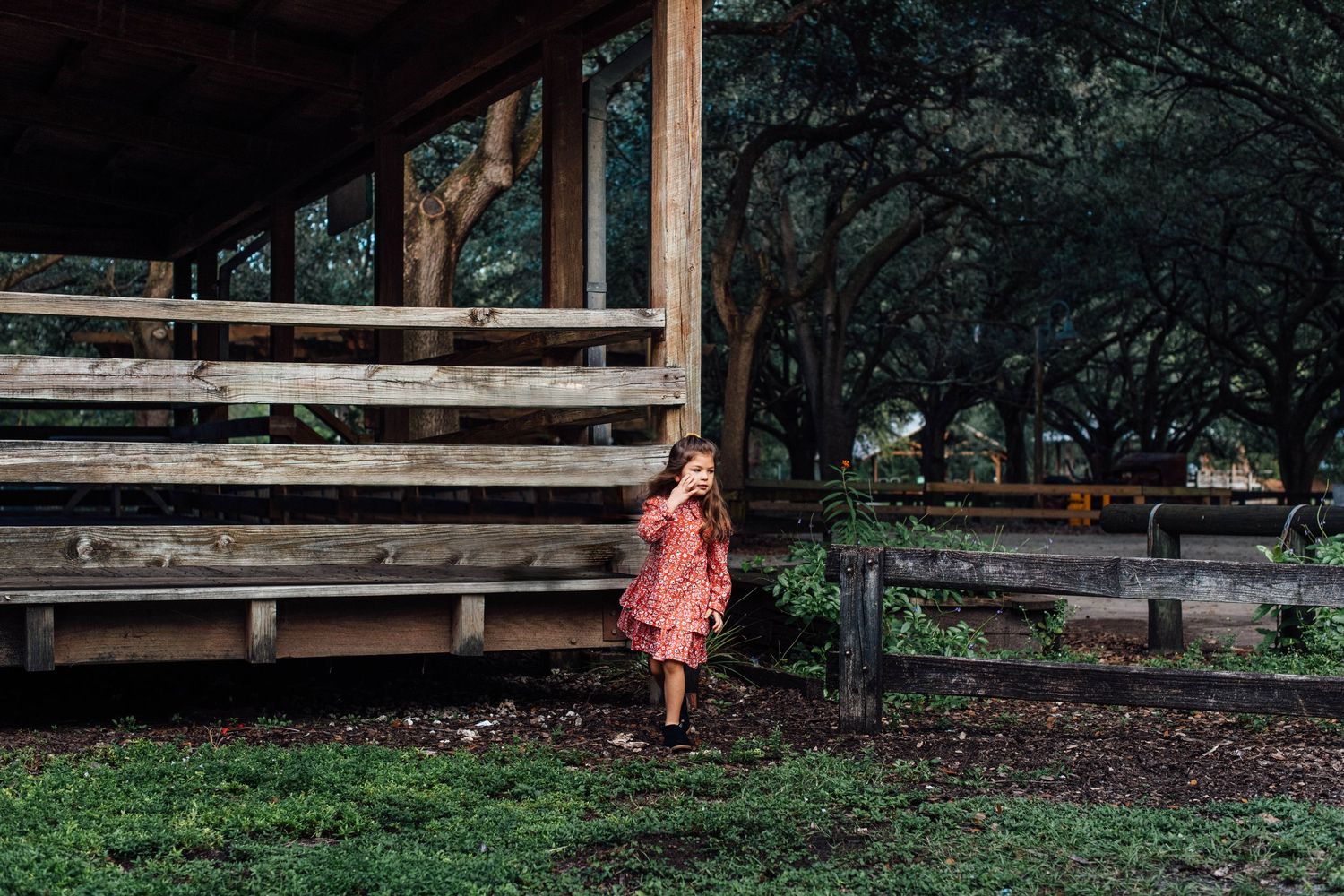 Young girl in red floral dress standing beside rustic wooden porch steps in a green park.