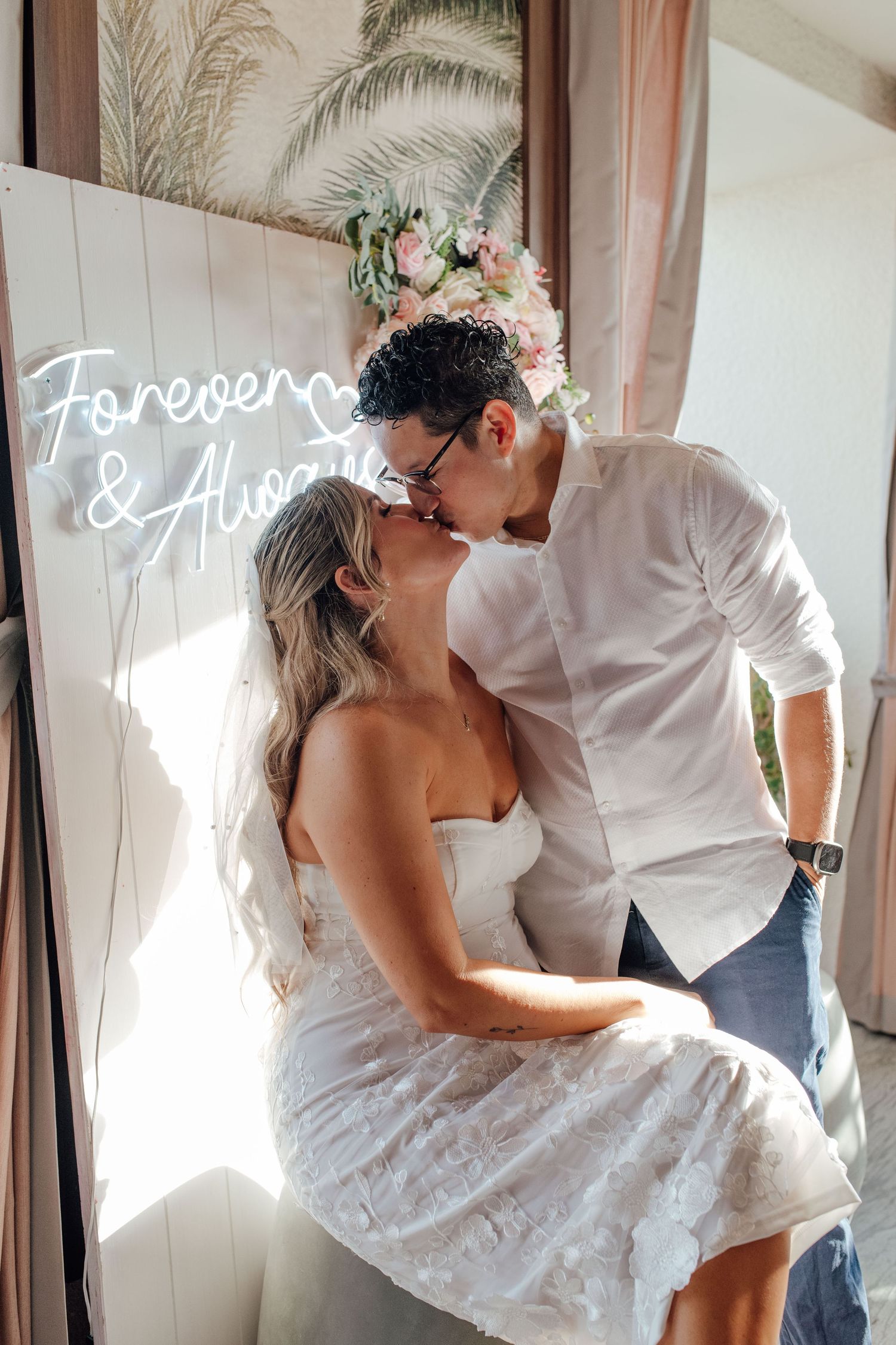 Bride and groom sharing a kiss in front of a Forever & Always neon sign at their wedding.