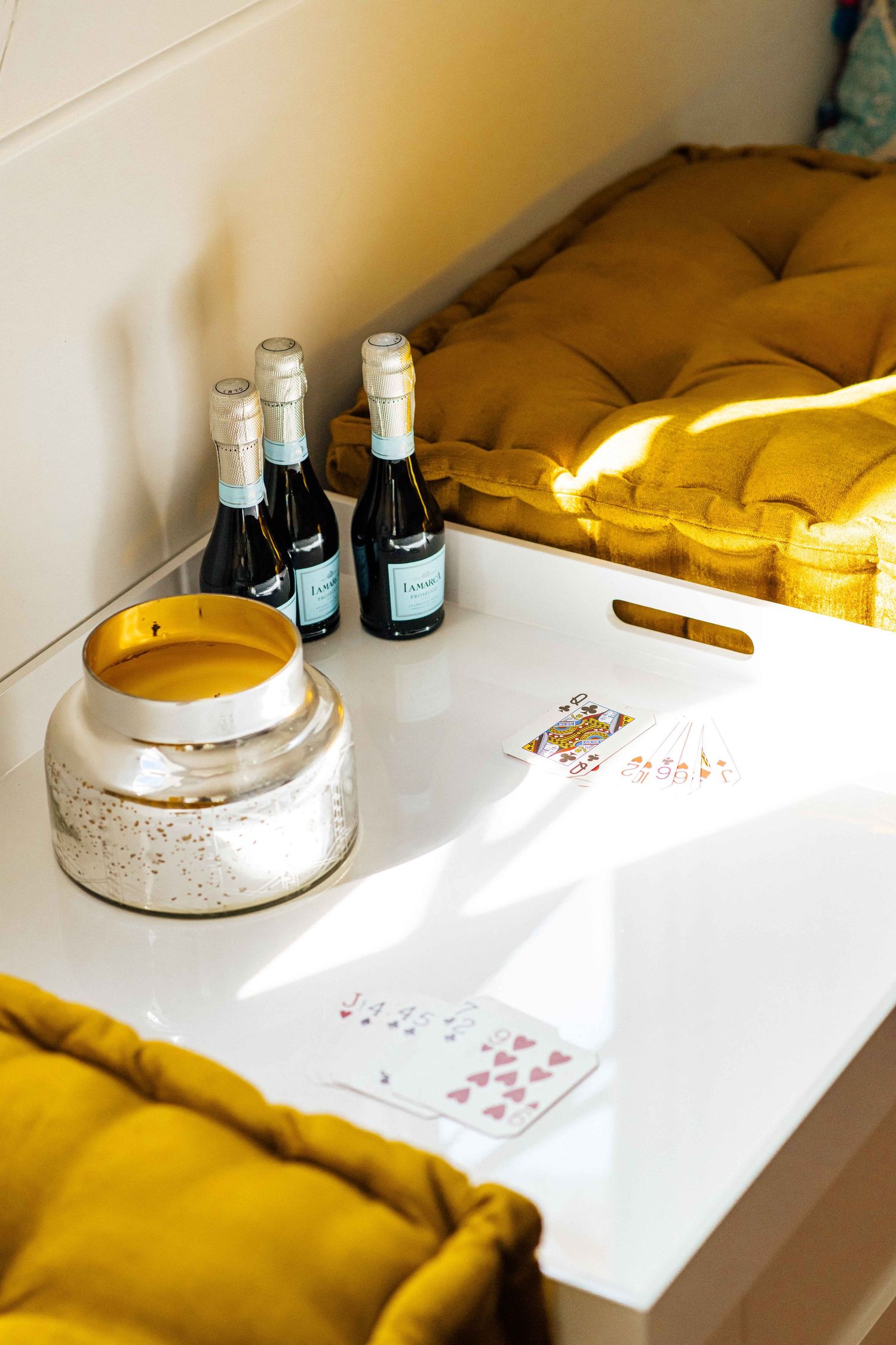 White tray with playing cards, candle jar, and mini champagne bottles beside mustard yellow cushions.