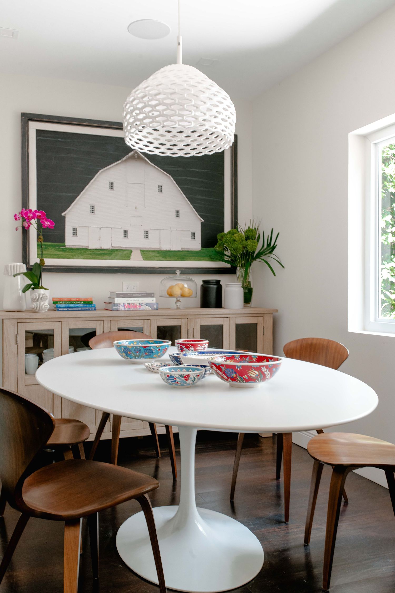 White tulip dining table with colorful bowls beneath a woven pendant light, wood chairs, and a barn painting on the wall.