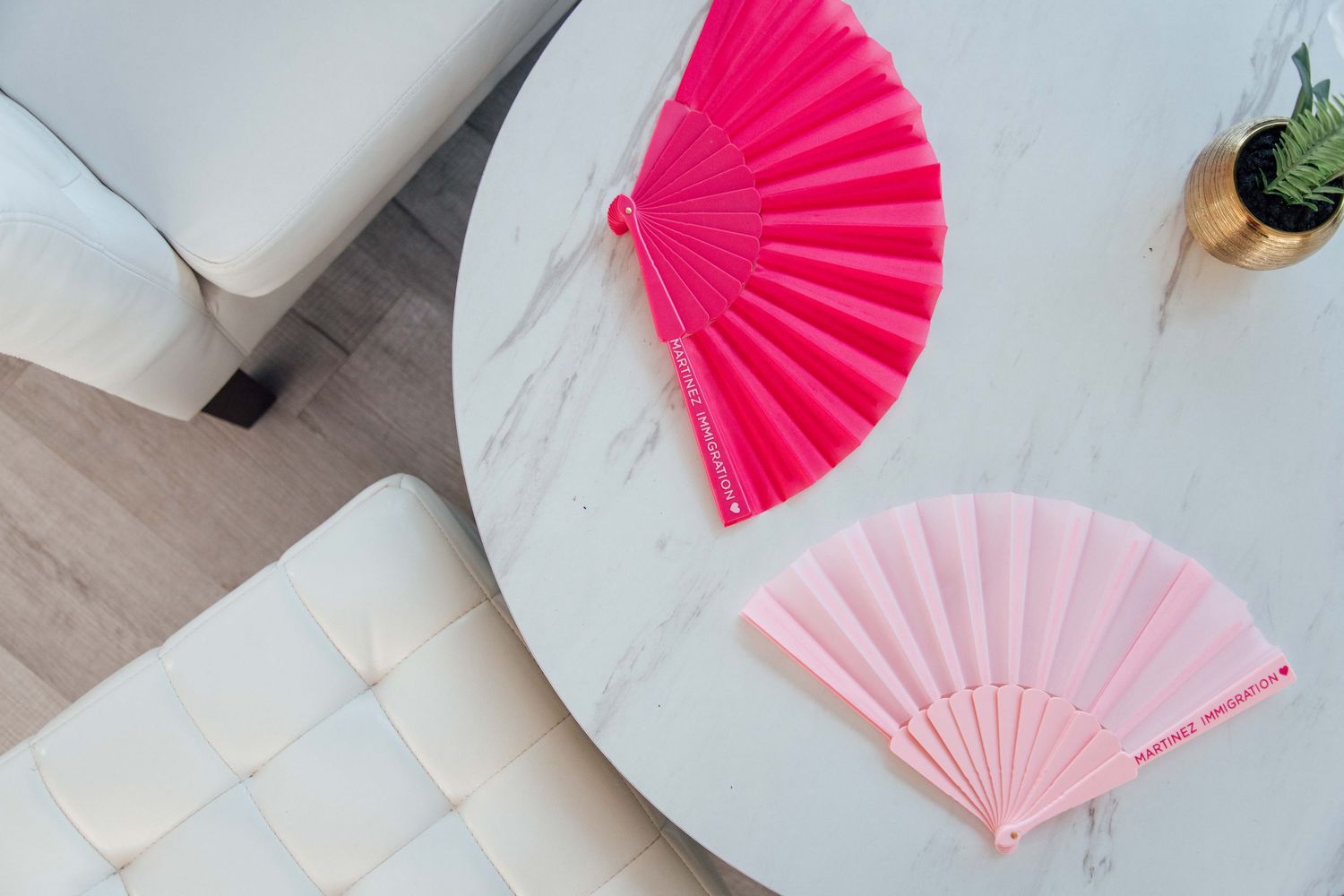 Two decorative hand fans in hot pink and light pink on a white marble table with a white sofa nearby.