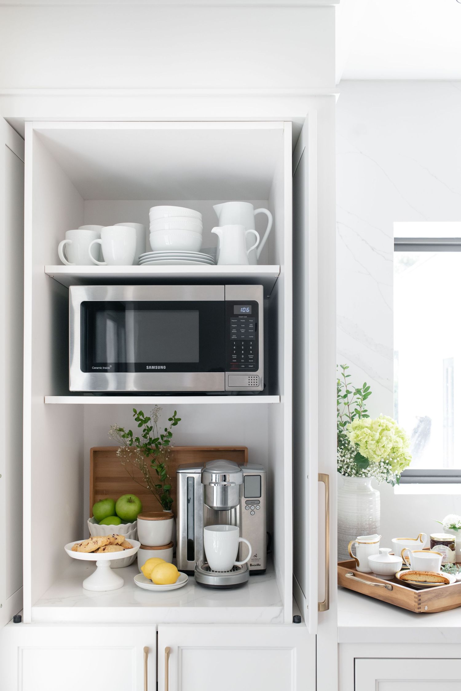White kitchen cabinet with Samsung microwave, Keurig coffee maker, green apples, lemon, and stacked white dishes on shelves.