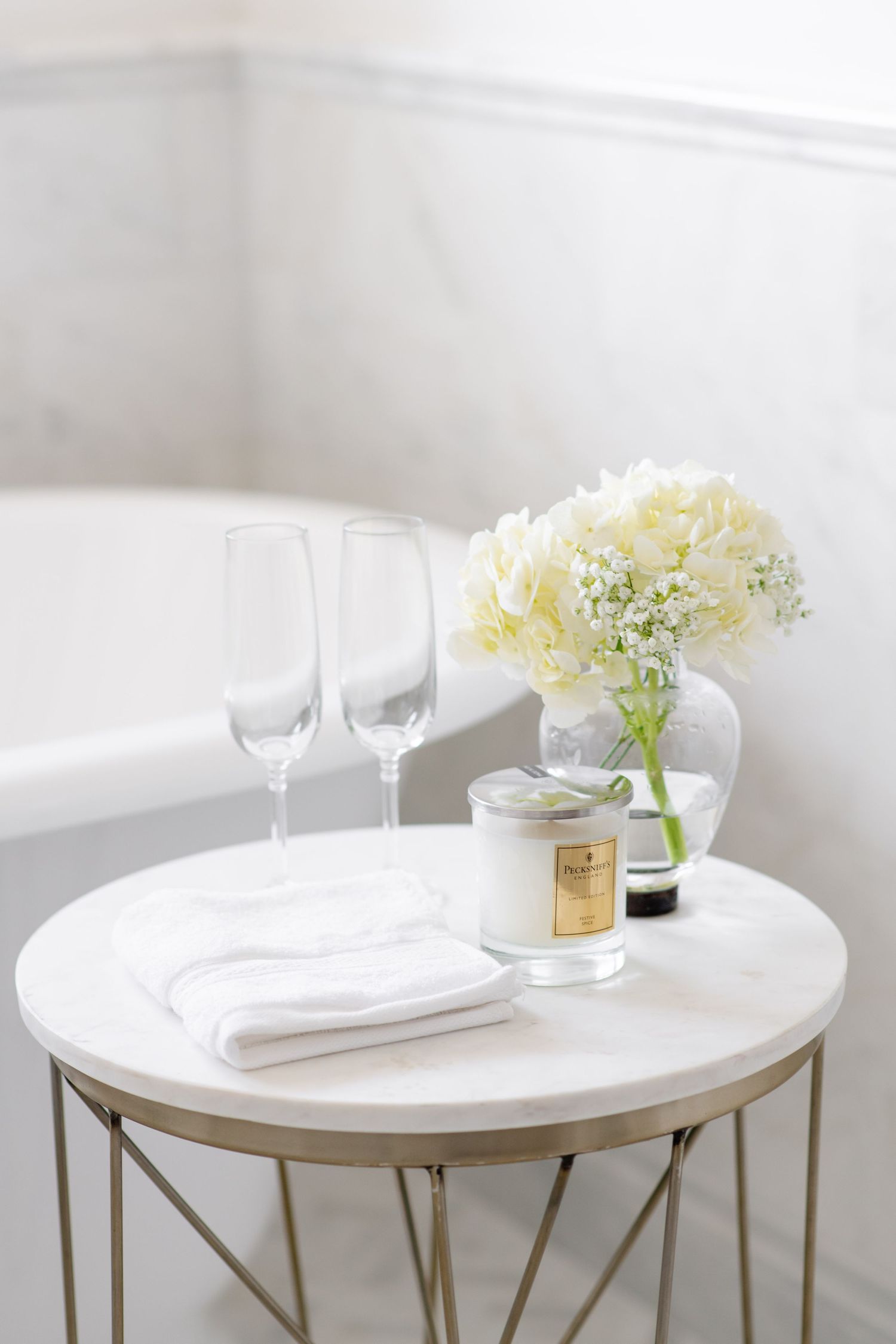 White marble side table with champagne flutes, folded towels, a scented candle, and white hydrangeas beside a bathtub.