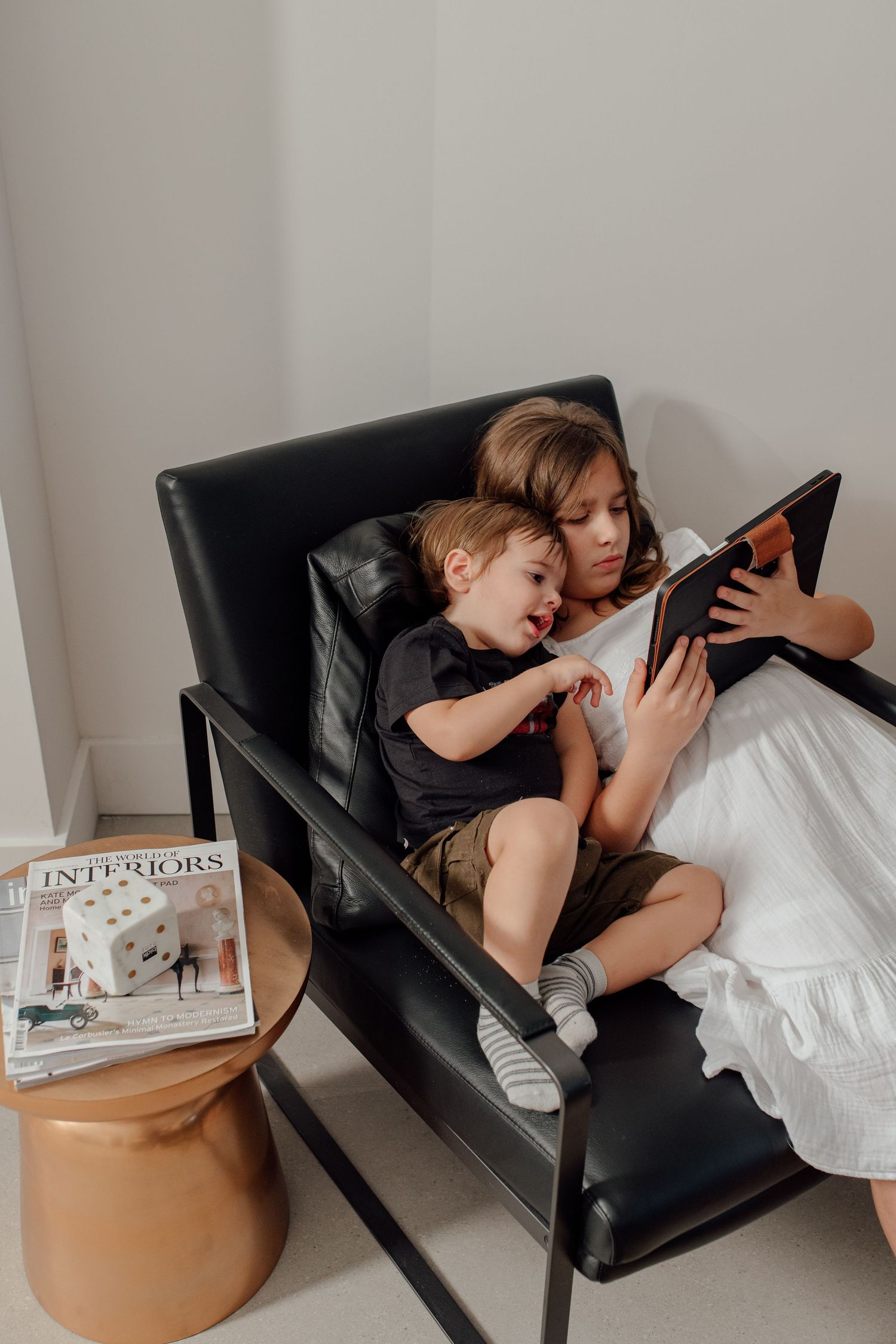 Two children sit together in a black leather chair, sharing a tablet, with an Interiors magazine on a copper side table nearby.