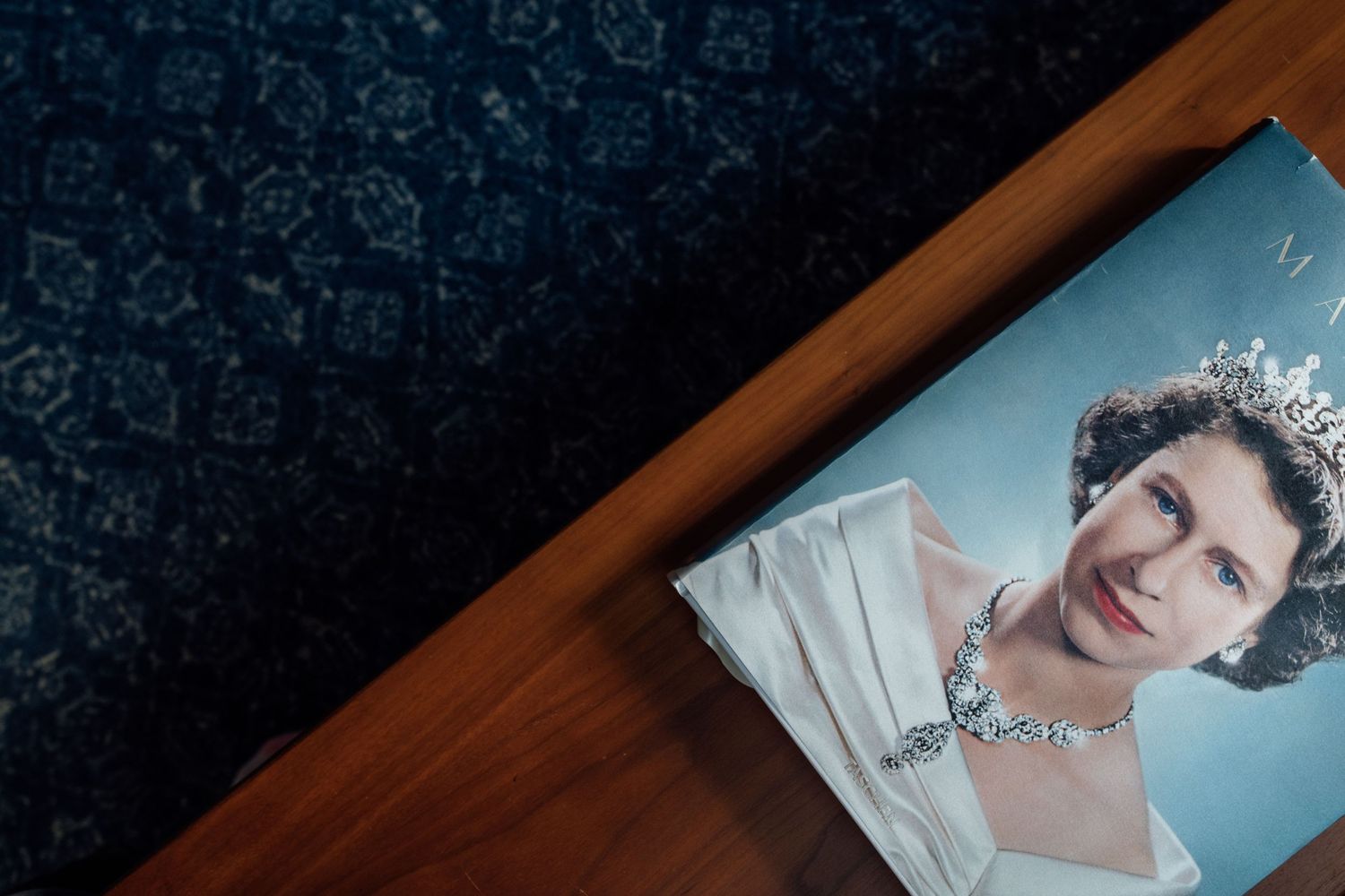 A portrait photograph of Queen Elizabeth II wearing a tiara and necklace, displayed on a wooden surface.
