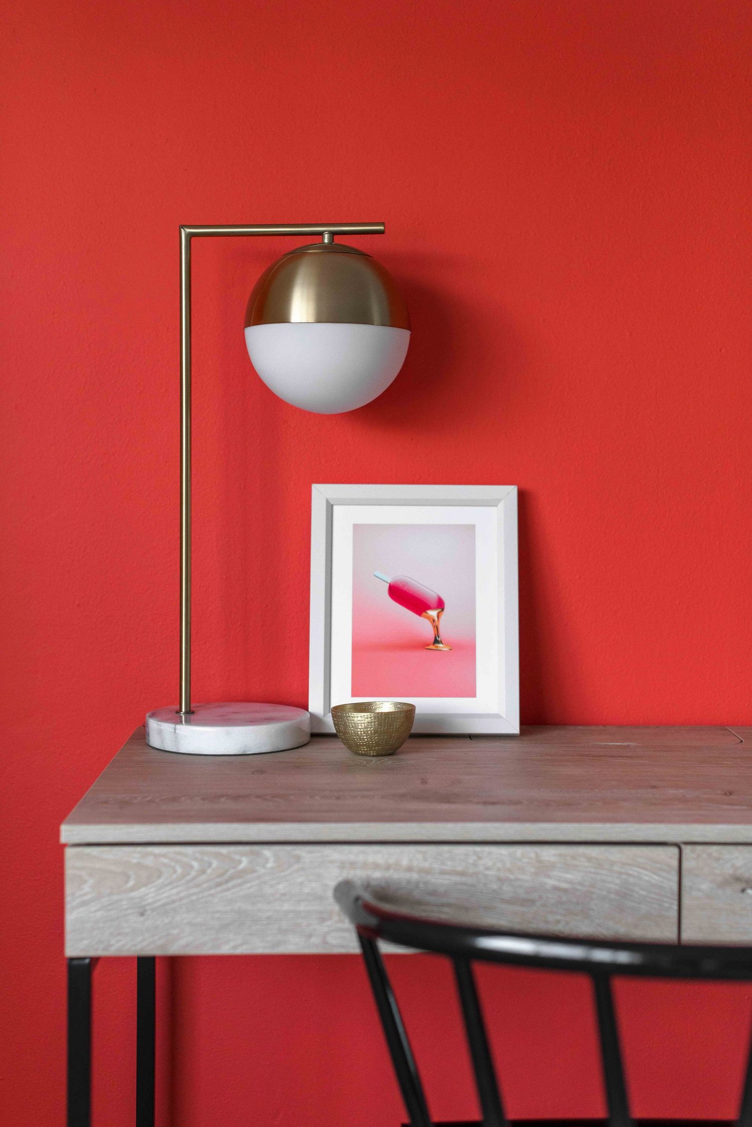 Modern desk setup with gold dome lamp, marble base, and framed art against bold red wall.
