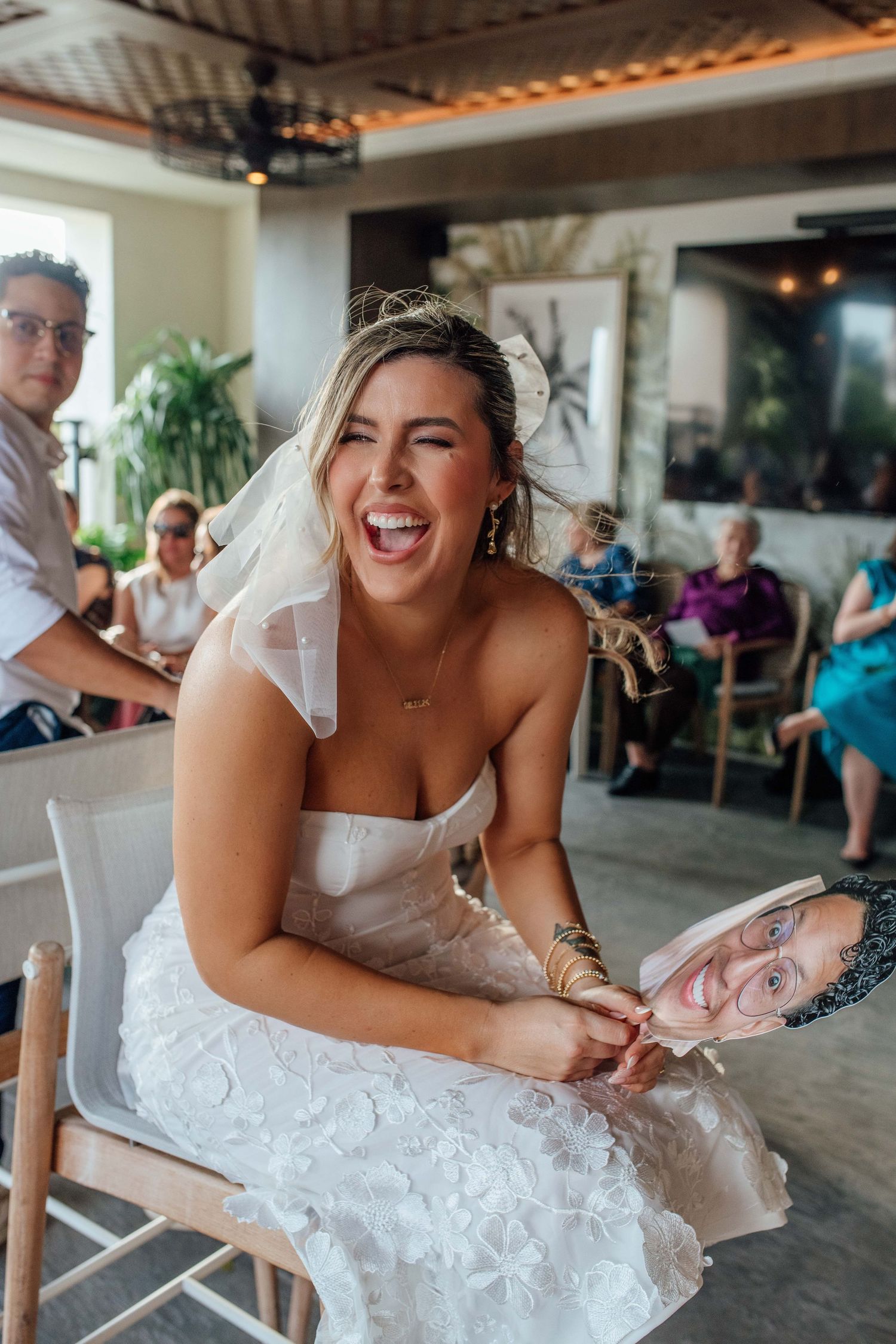 Laughing bride in strapless white gown holds a face cutout prop during wedding reception celebration.