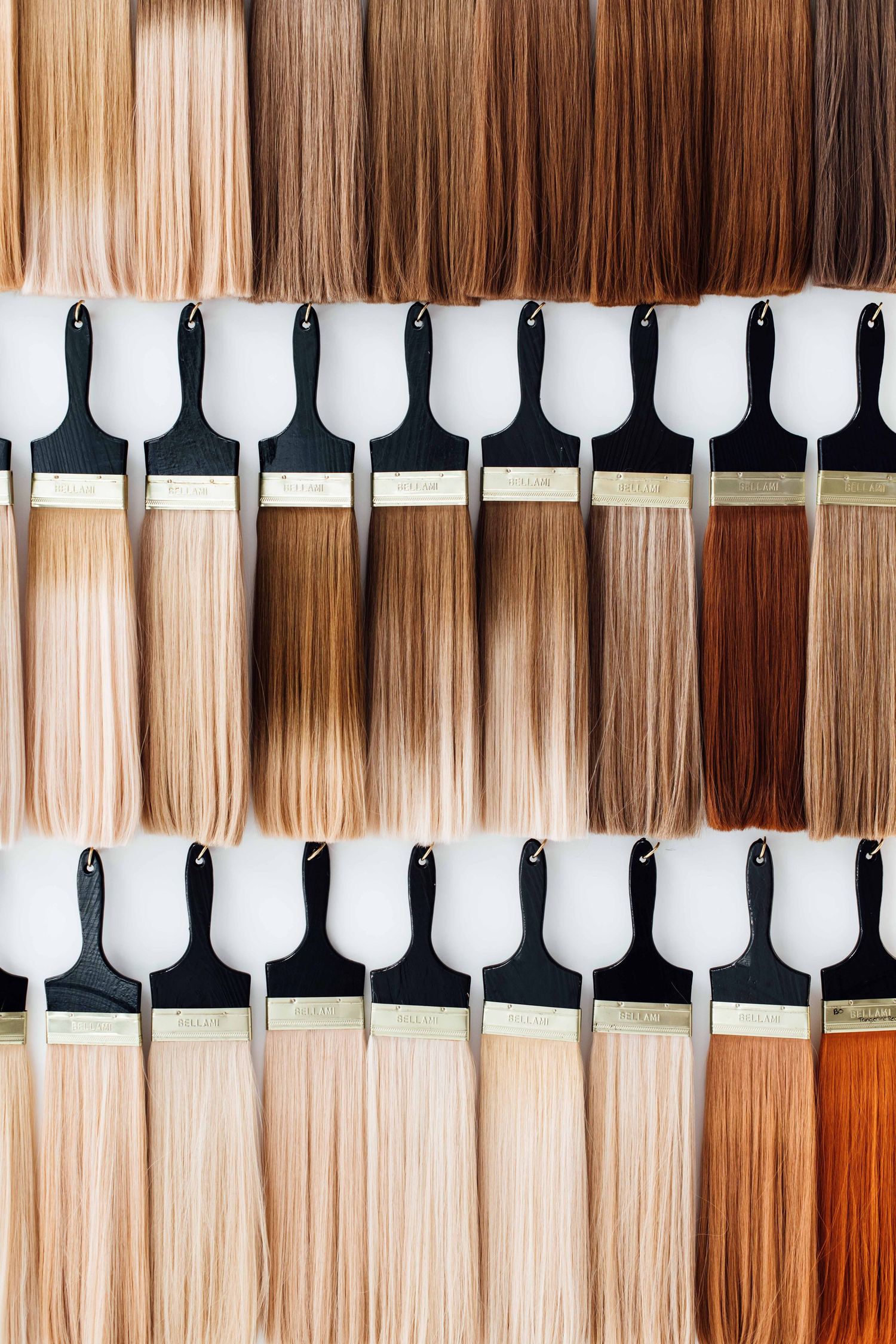 A hair color swatch display showing rows of straight hair extensions ranging from blonde to dark brown shades on white brushes.