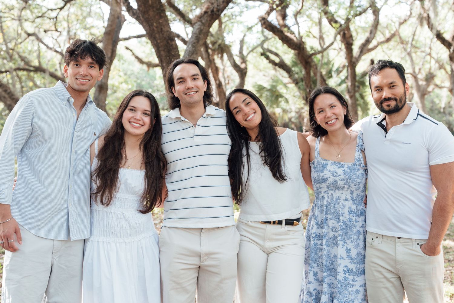 Six smiling family members pose together outdoors under large oak trees wearing light-colored casual outfits.