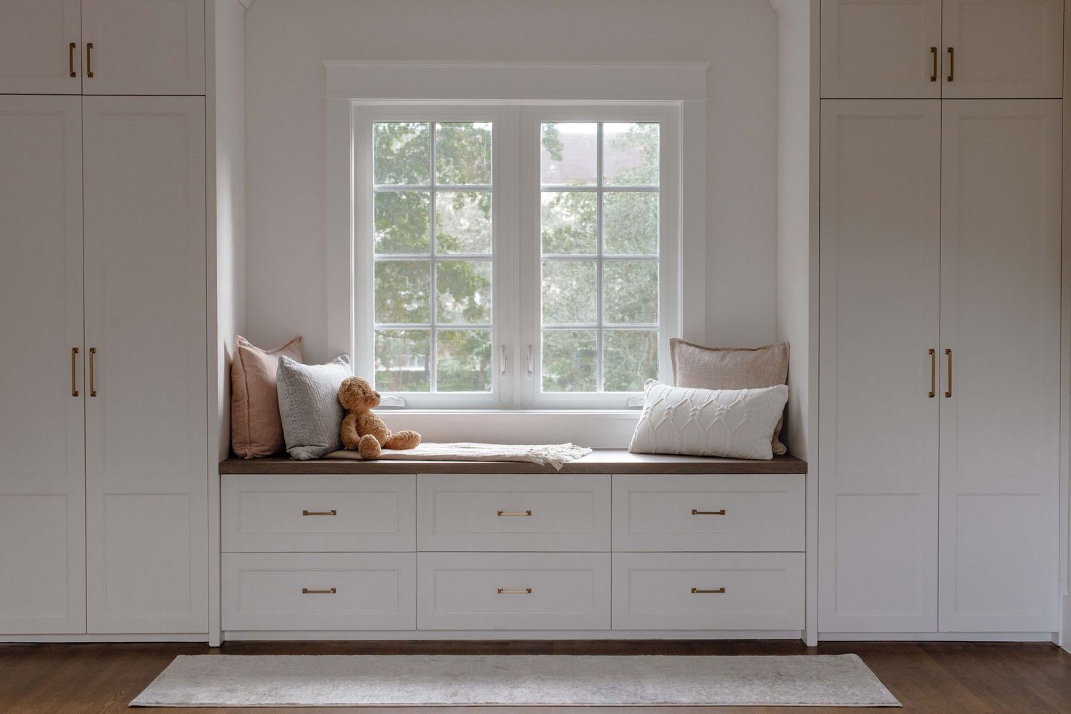 White built-in cabinets flank a cozy window seat with cushions and a teddy bear, featuring gold hardware and storage drawers.