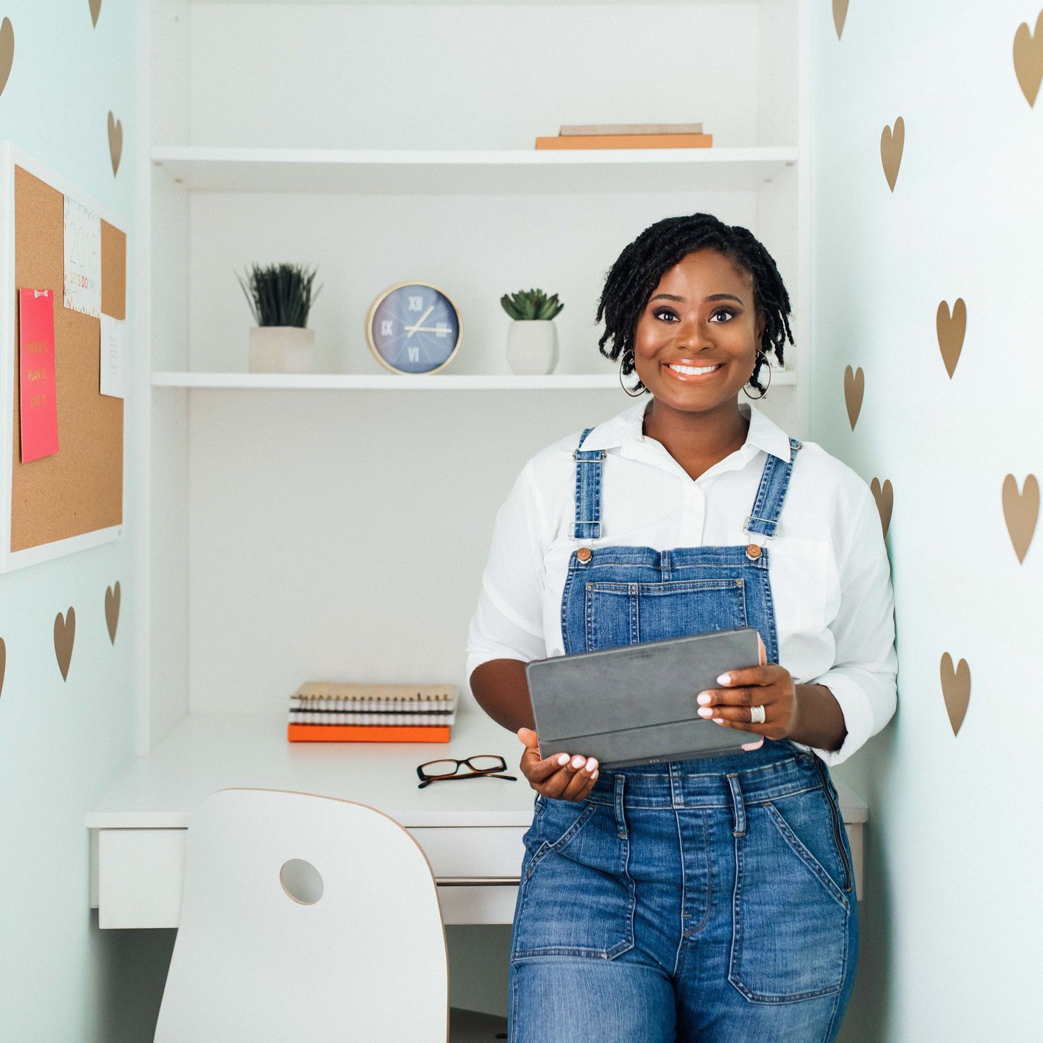Smiling Black woman in denim overalls holding a tablet in a decorated home office workspace.