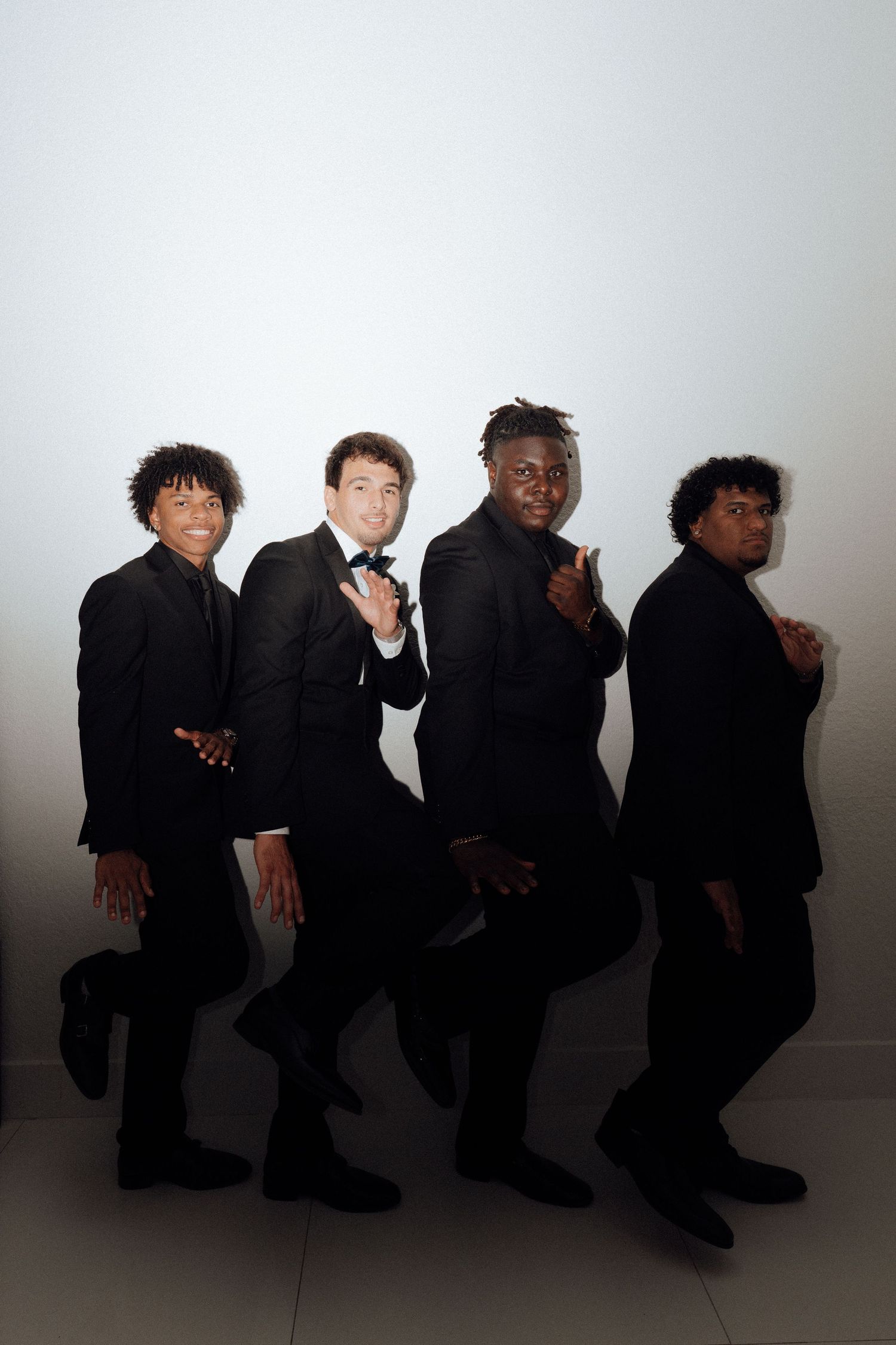 Four men in black tuxedos pose confidently against a white background in a stylish group photo.