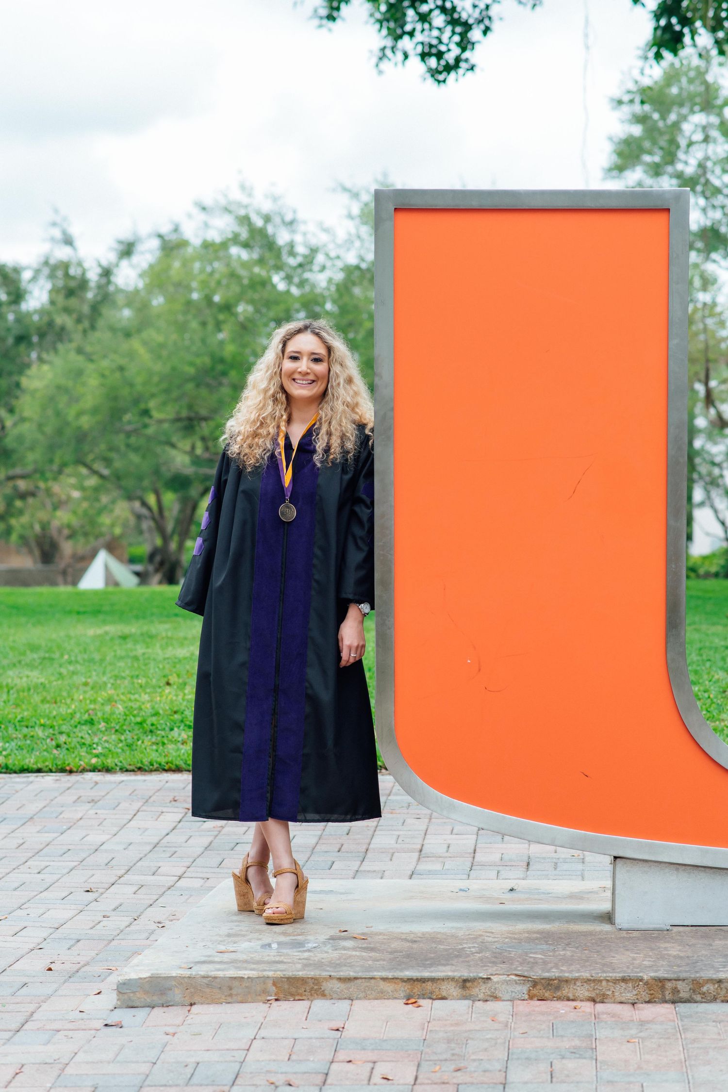 Smiling graduate in black gown and medal poses beside large orange university letter monument on brick campus path.