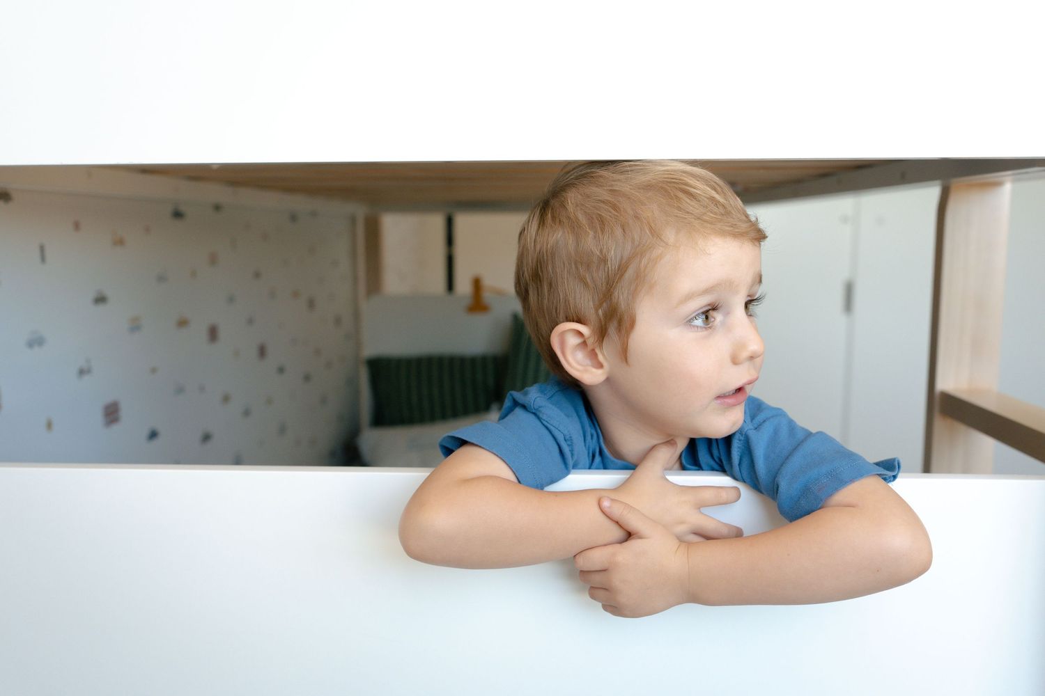 Curious young boy in blue shirt resting arms on white table, looking sideways in a bright modern room.