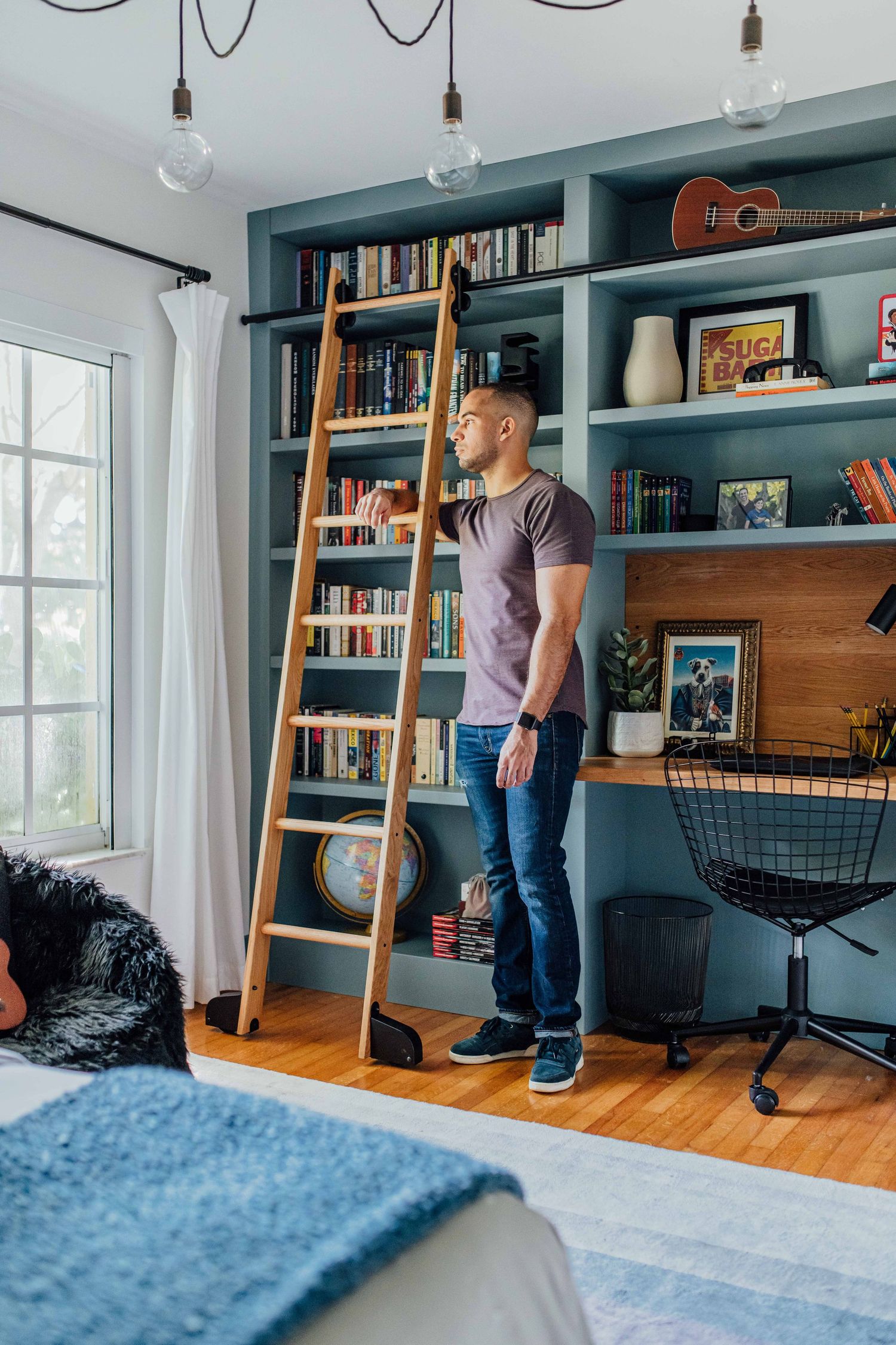 Man standing beside a wooden library ladder leaning against a built-in blue bookshelf with desk in a modern bedroom.