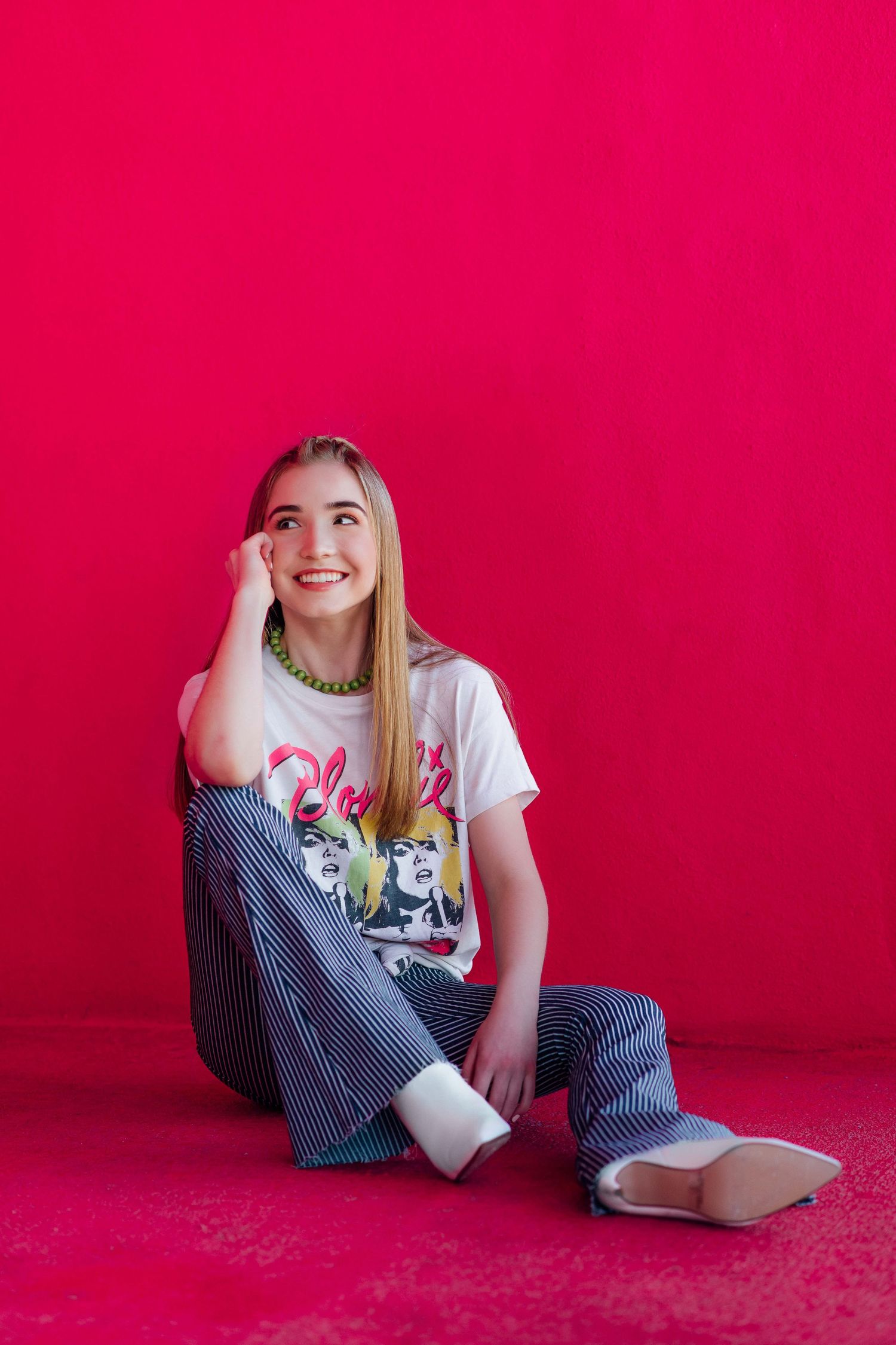 Smiling teenage girl in a graphic tee and striped pants sitting on a pink floor against a red wall.