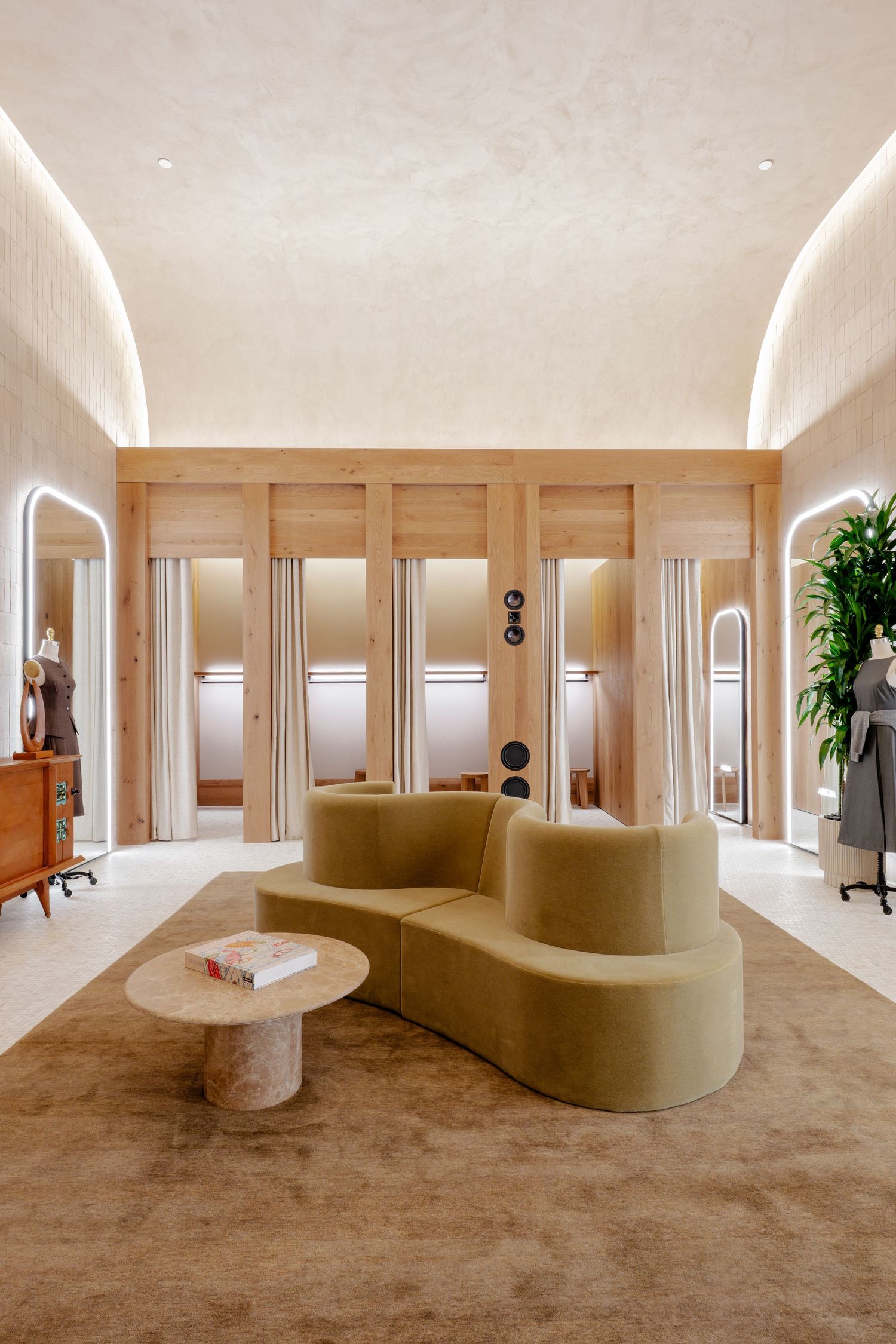 Elegant boutique interior with curved olive green sofa, stone coffee table, and warm wood fitting rooms.