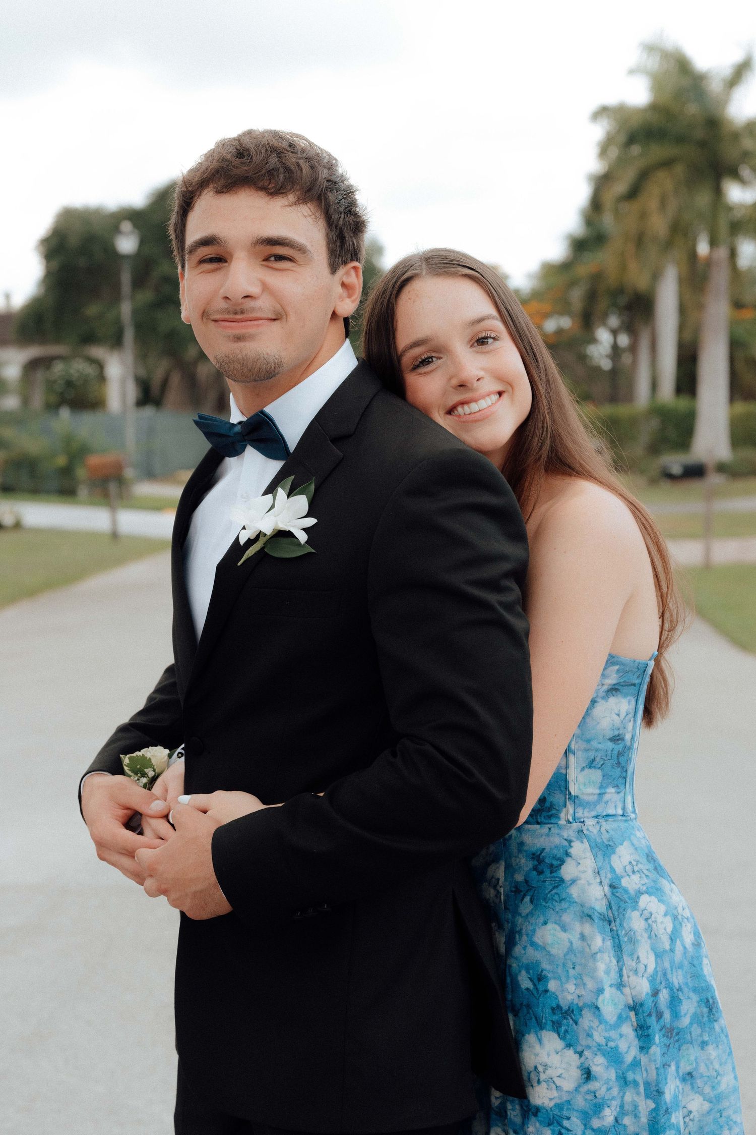 Young couple dressed for prom, man in black tuxedo with boutonniere, woman in blue floral dress, posing outdoors.