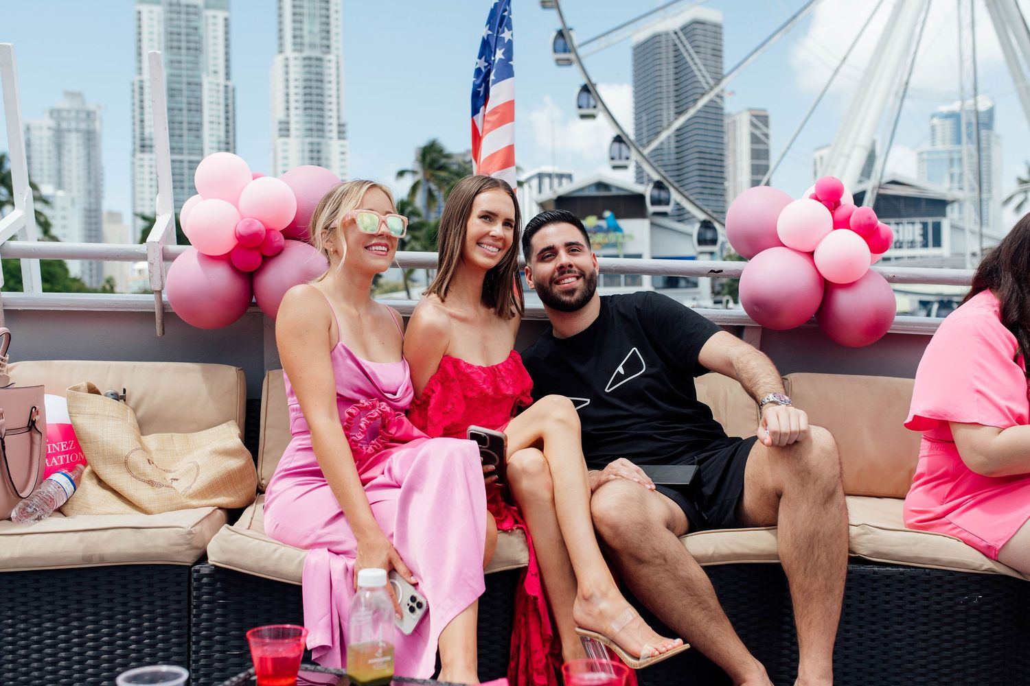 Two women in pink dresses and a man sit together at an outdoor waterfront party with pink balloons.