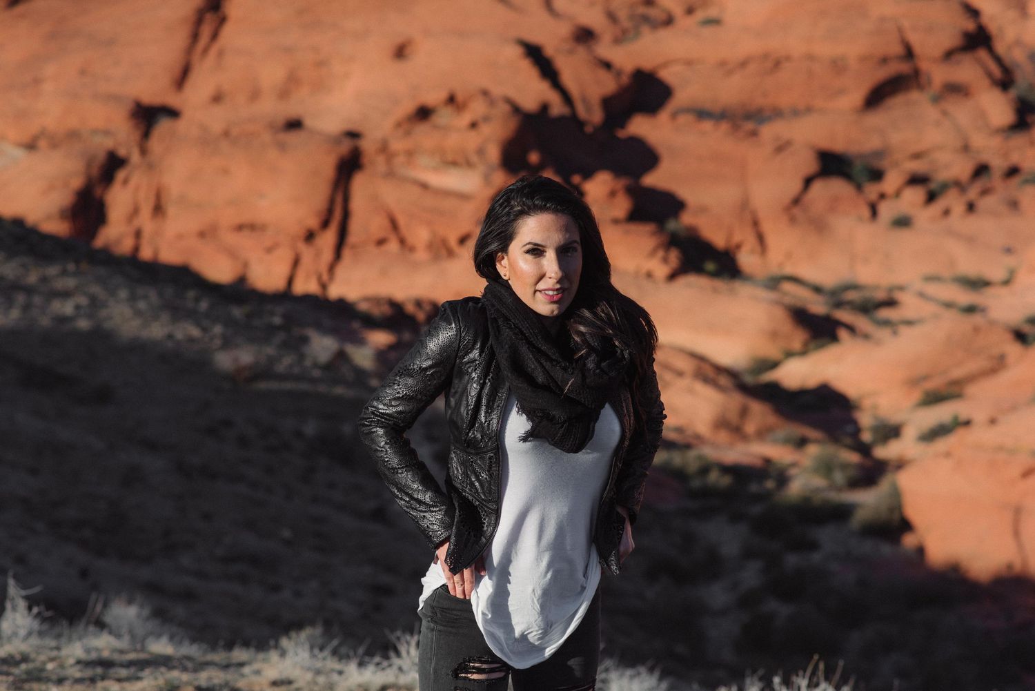 Woman in leather jacket and white top posing before red rock formations in natural desert landscape.