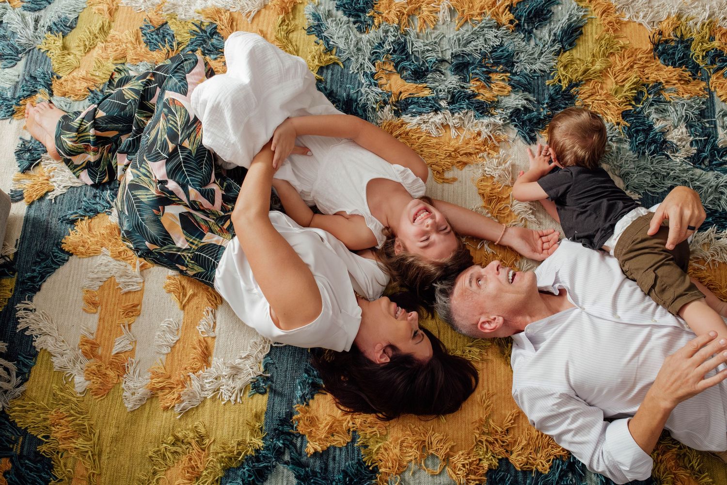 Family of four lying on a colorful floral rug, parents smiling while holding their two young children.