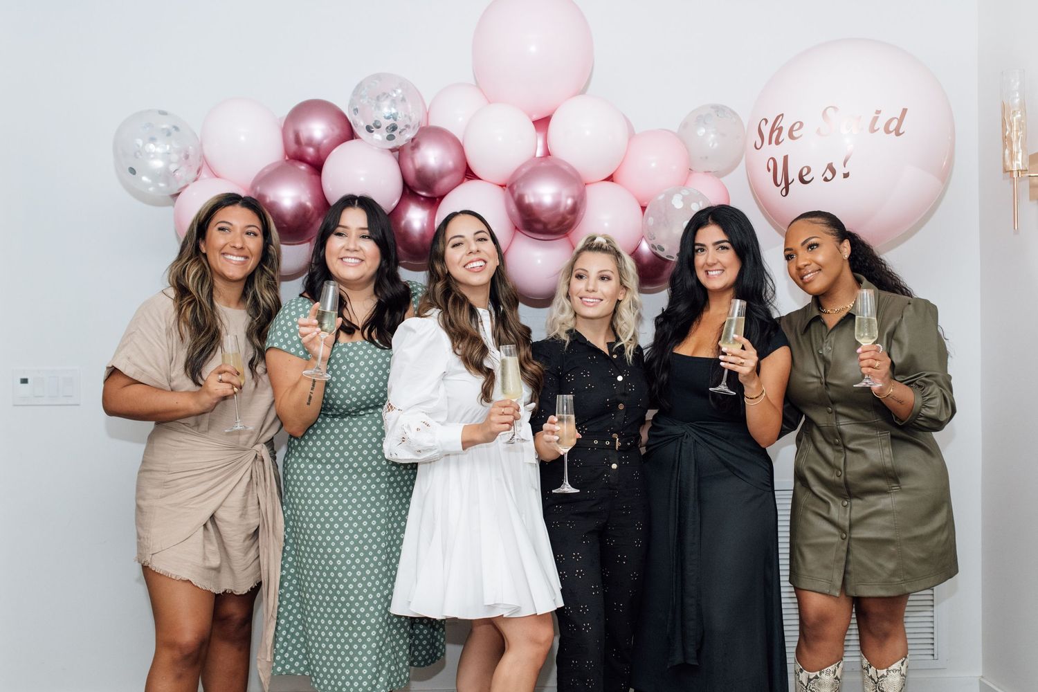 Six women celebrating a bachelorette party with champagne in front of pink balloon wall decoration.