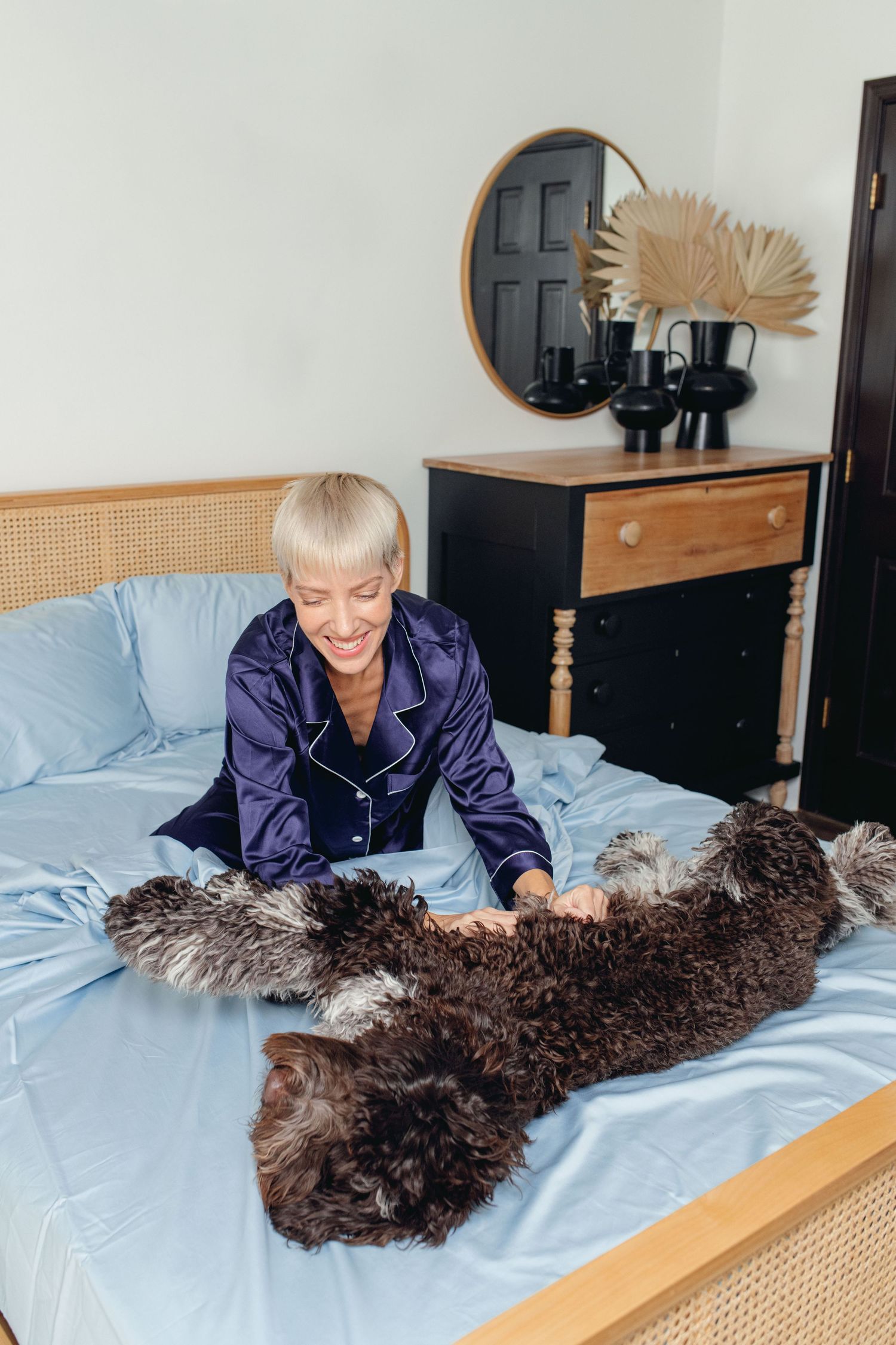 Smiling blonde woman in purple pajamas petting a large dark curly dog on blue bedding.