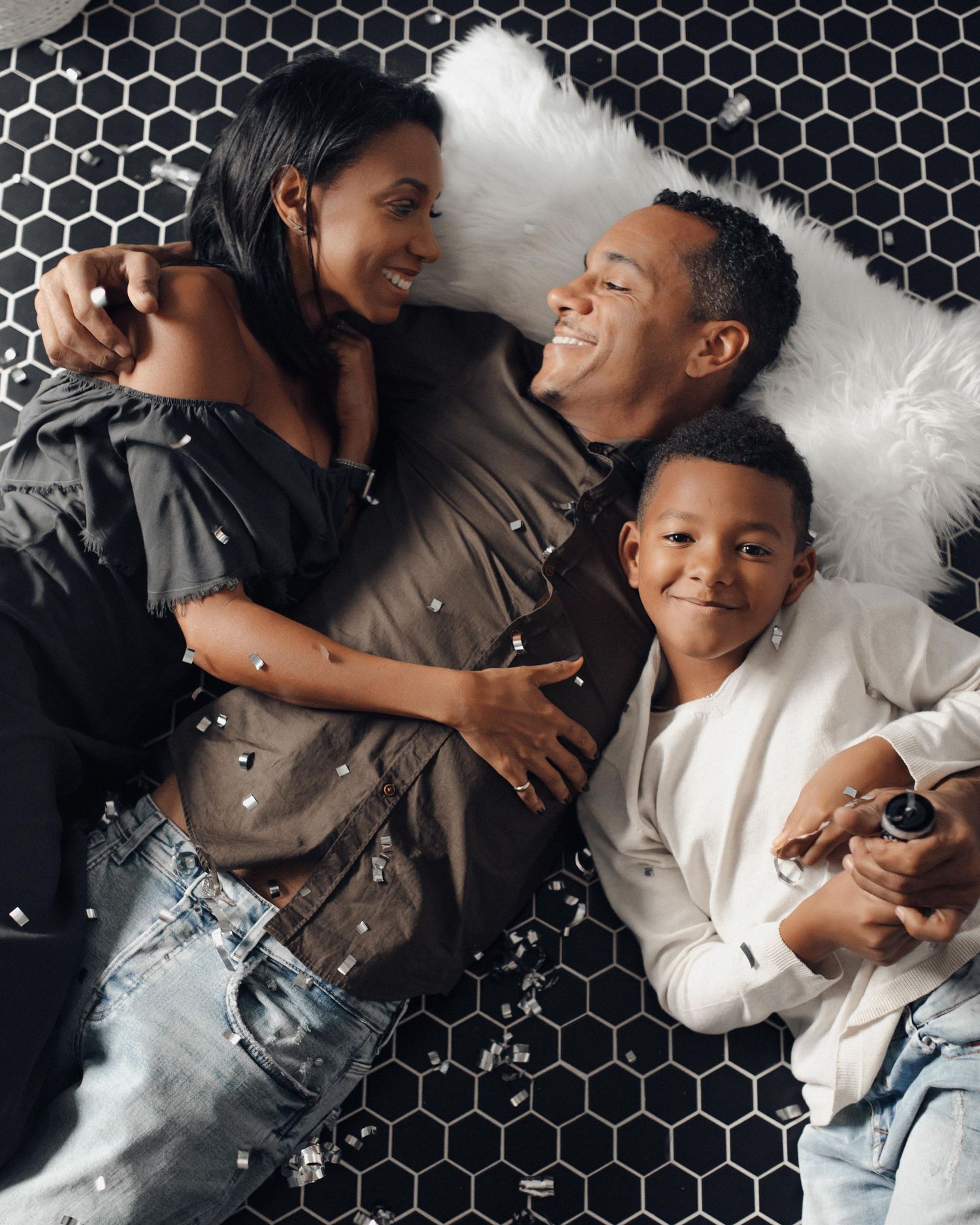 Happy family of three lying on black hexagon tile floor, smiling and embracing each other.