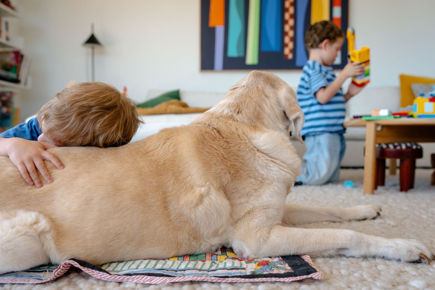 Young boy hugging a golden retriever dog on carpet while another child plays with toys in background.