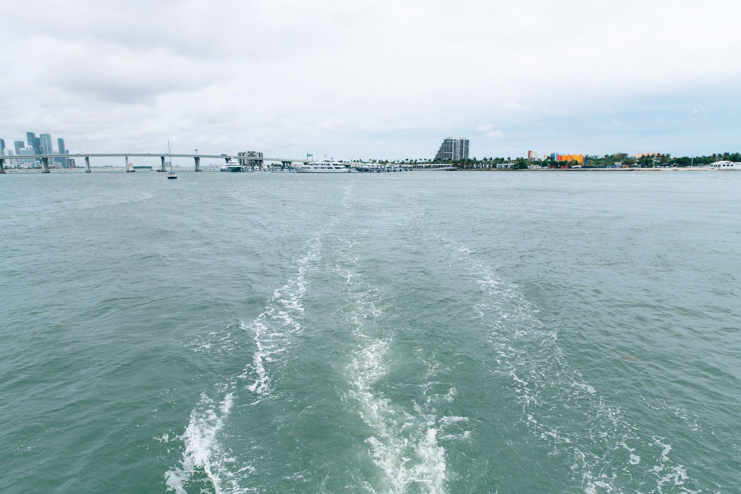 Boat wake trail on calm ocean waters with a city skyline and bridge visible in the background under cloudy skies.