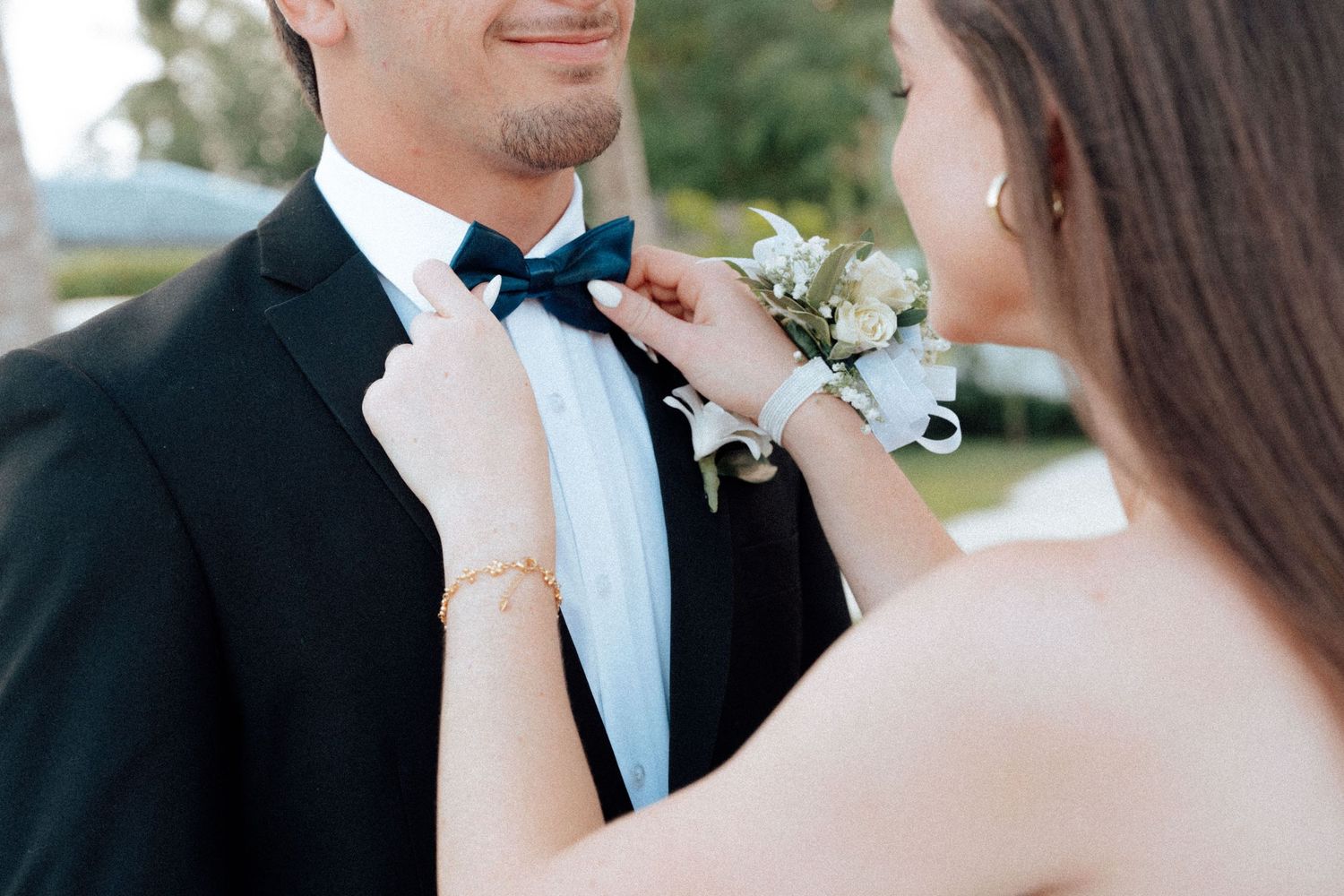 Woman pinning a white boutonniere on a man in a black tuxedo with a navy bow tie at prom.