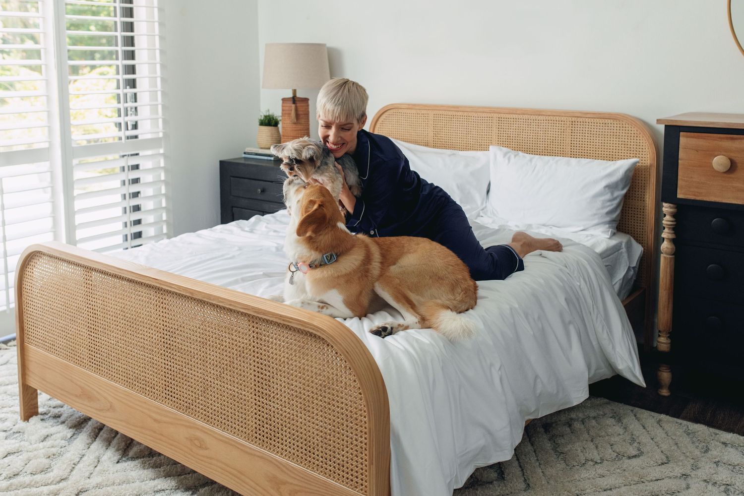 Person cuddling with a golden retriever dog on a rattan bed with white bedding near window.