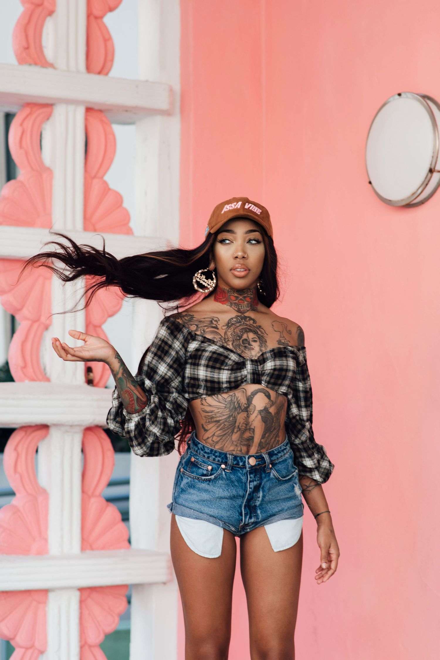 Tattooed woman in plaid crop top and denim shorts poses against pink wall with white decorative trim.