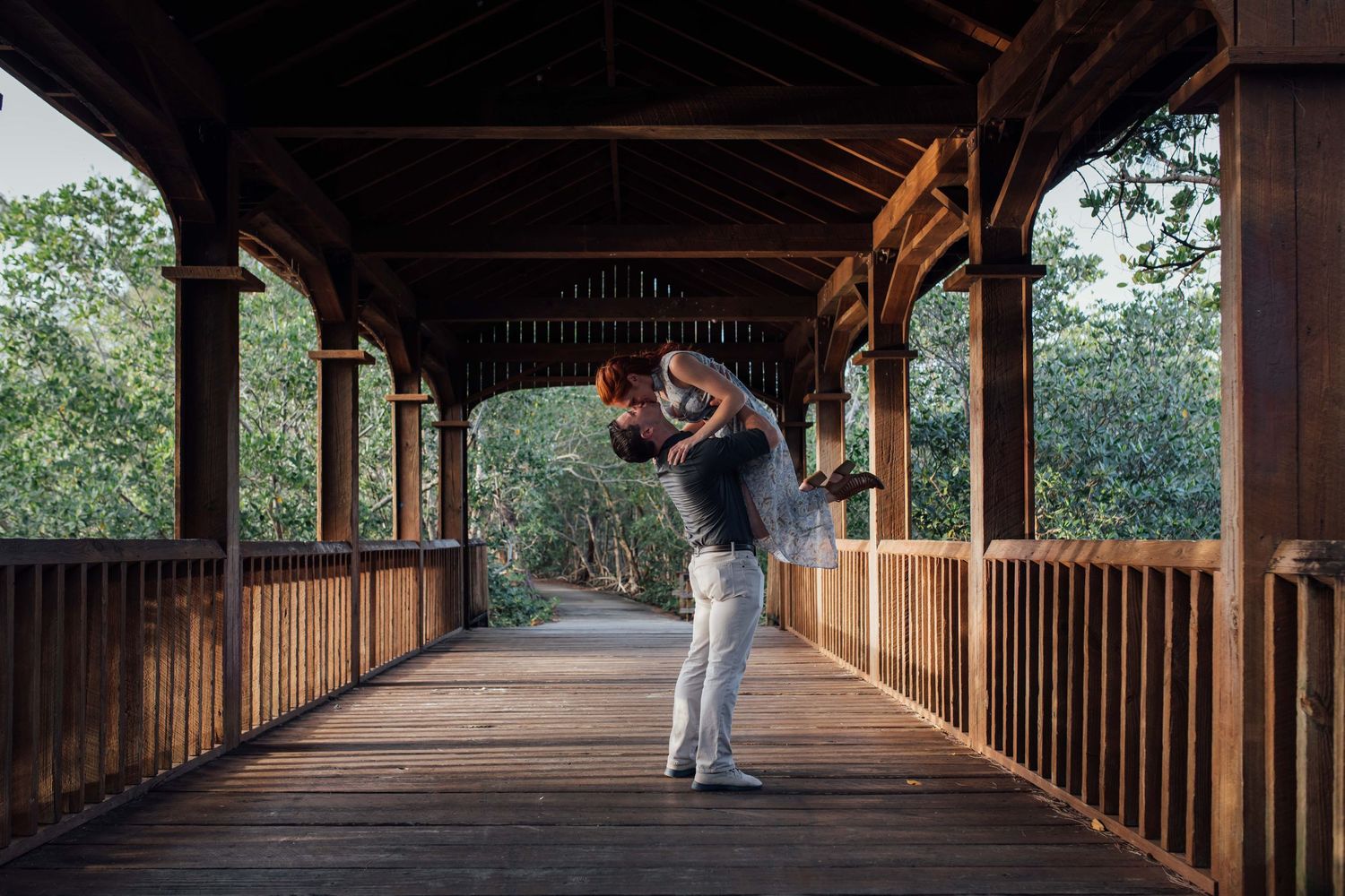 Couple sharing a romantic embrace on a wooden covered bridge surrounded by lush green trees.