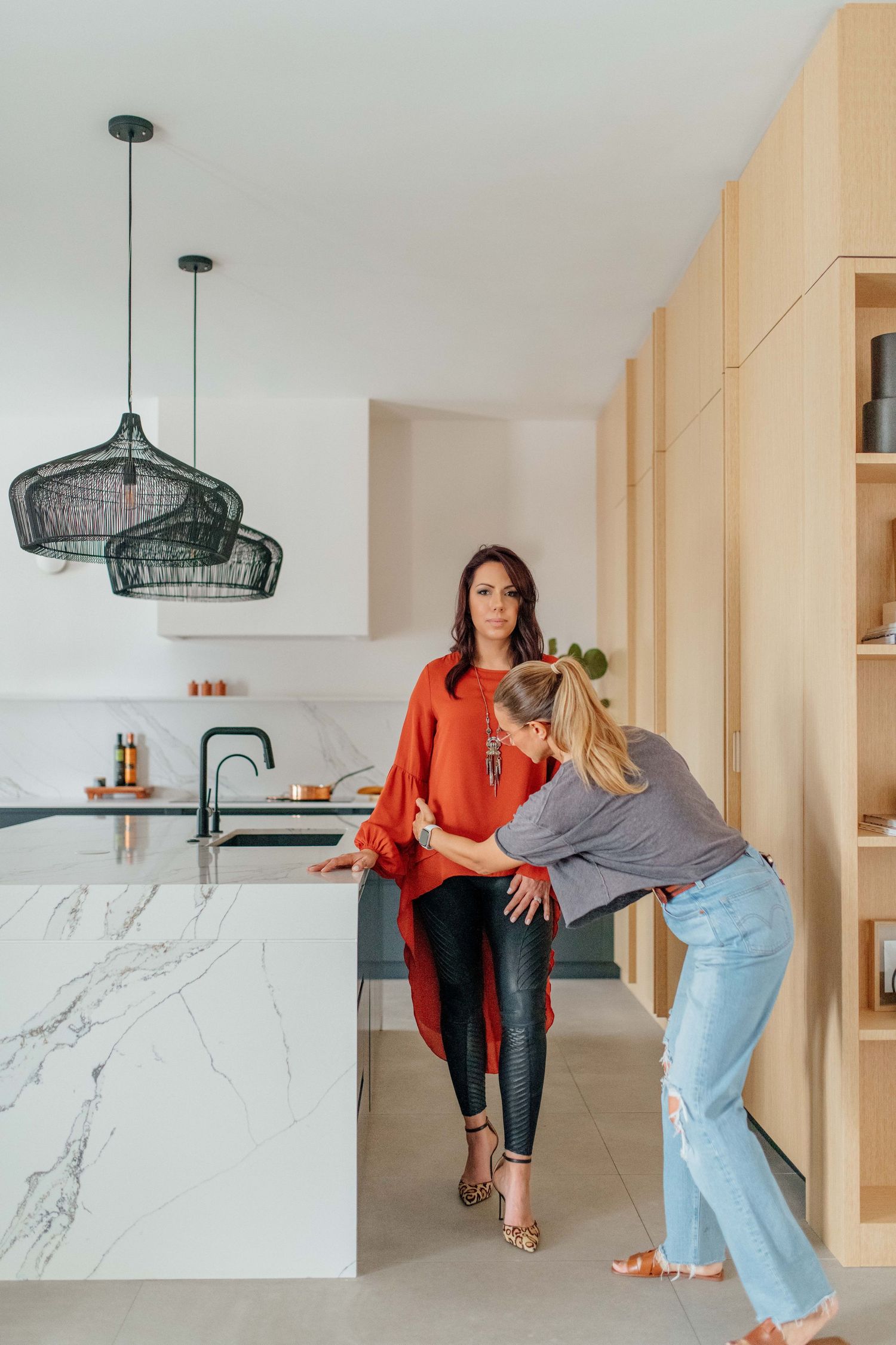Two women in a modern kitchen with marble island, pendant lights, and wood cabinetry, one in red top, one in jeans.