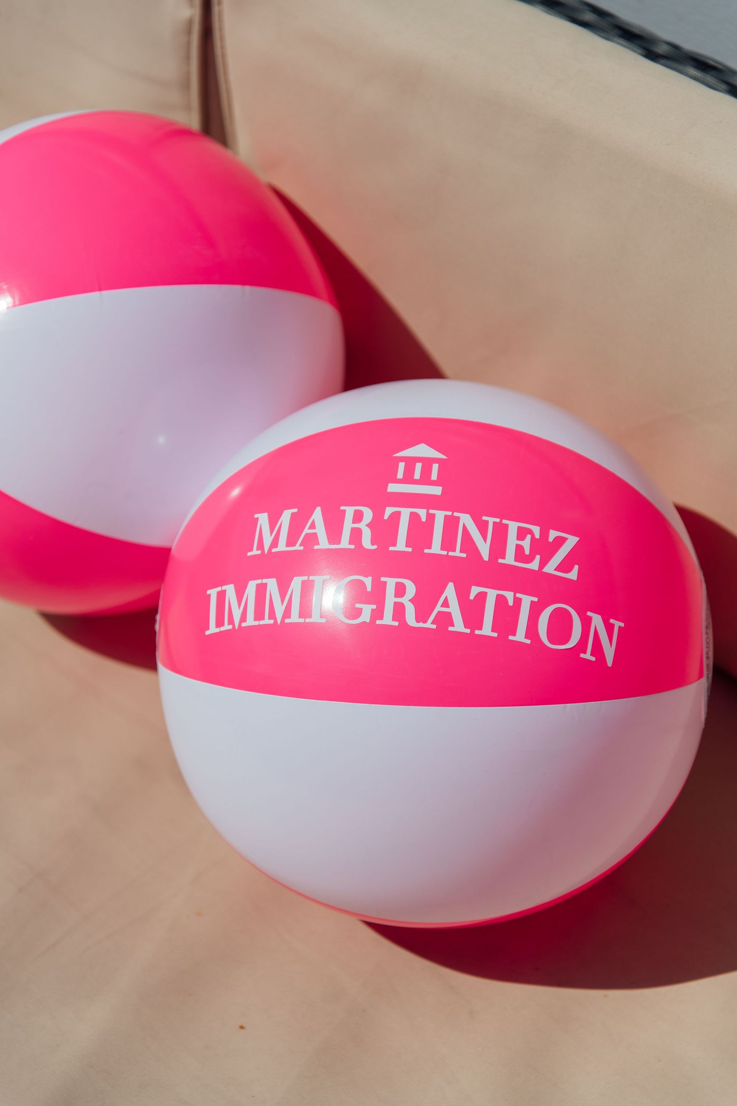 Pink and white inflatable beach balls printed with Martinez Immigration logo and building icon on sandy surface.