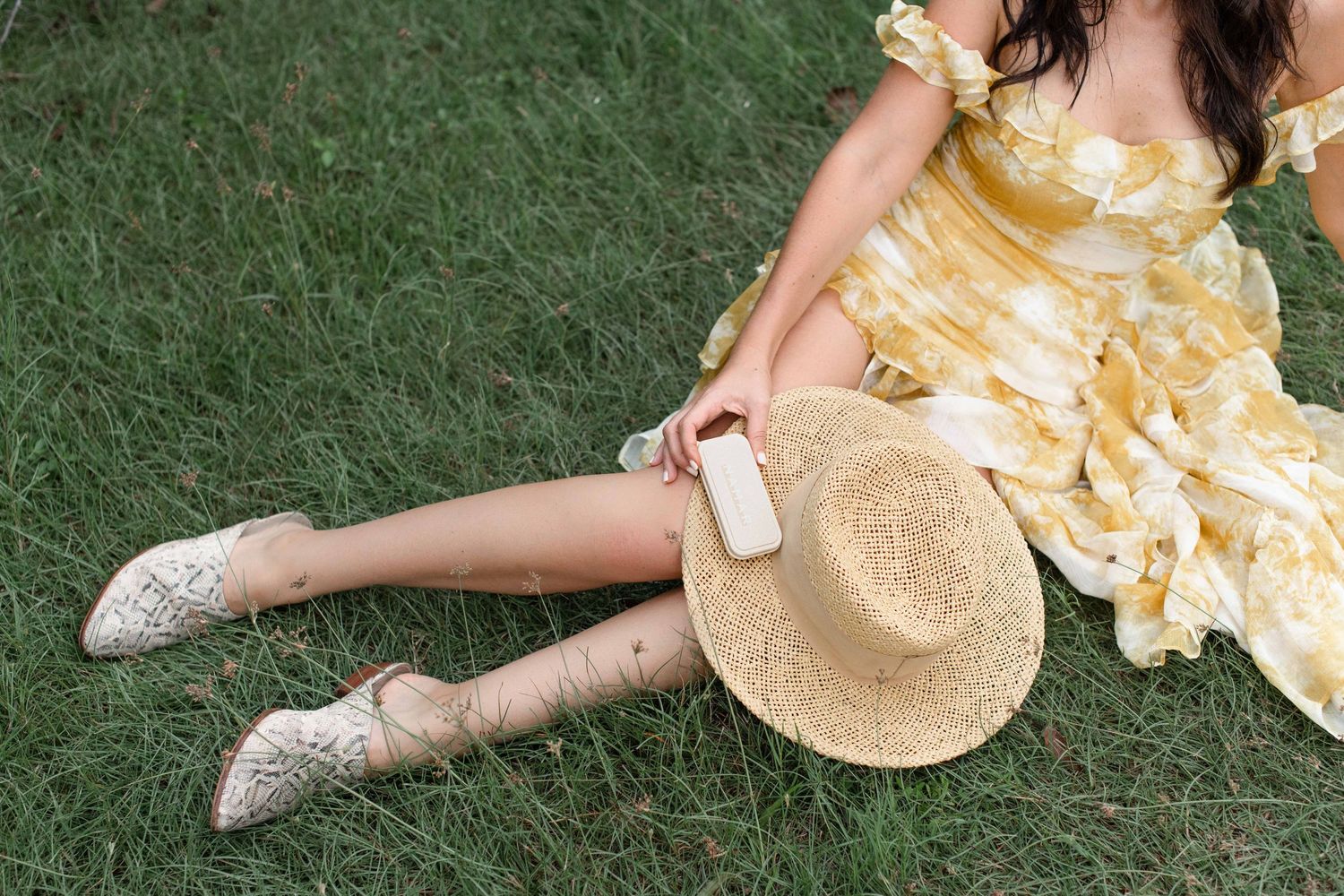 Woman in yellow floral dress sitting on green grass holding phone with straw hat beside her.