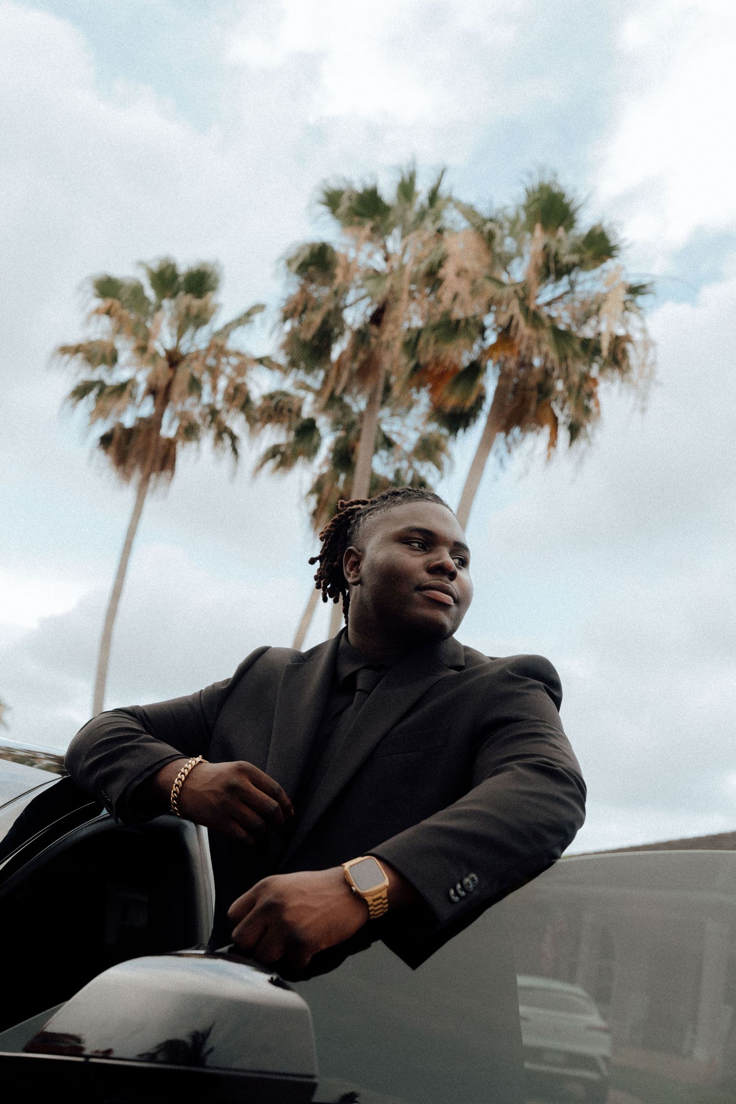 A stylish young Black man in a black suit with gold jewelry leans on a car, palm trees and cloudy sky behind him.