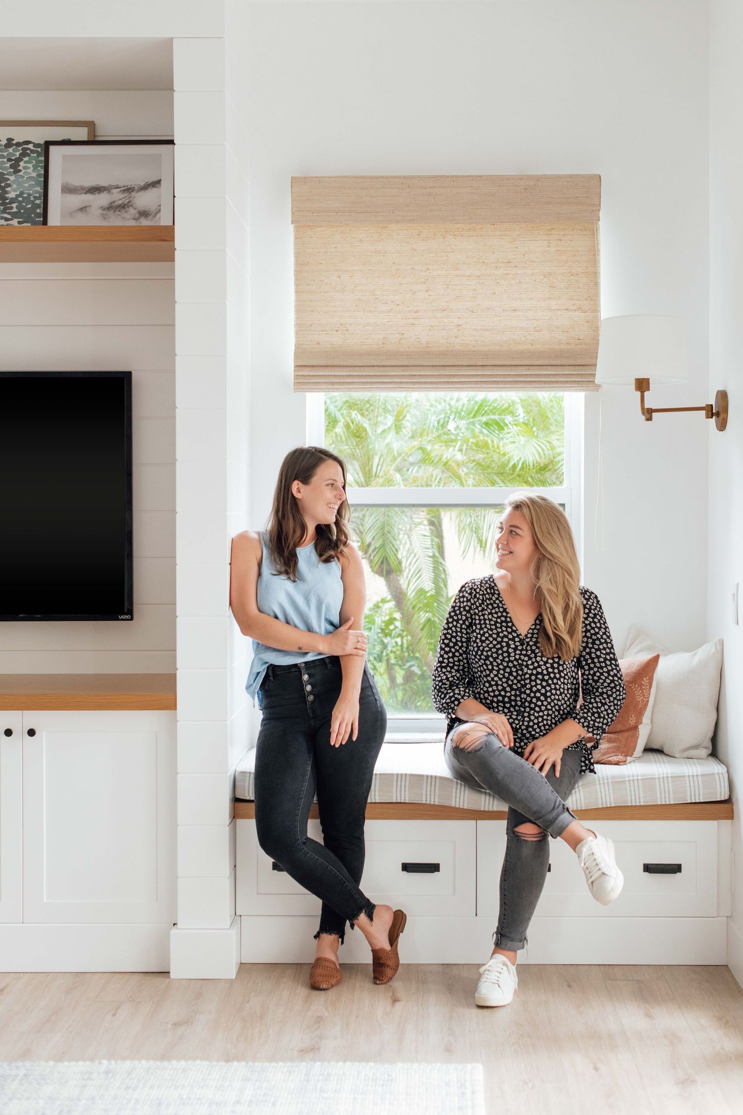 Two women laughing together in a bright white living room with built-in shelving and window seat.