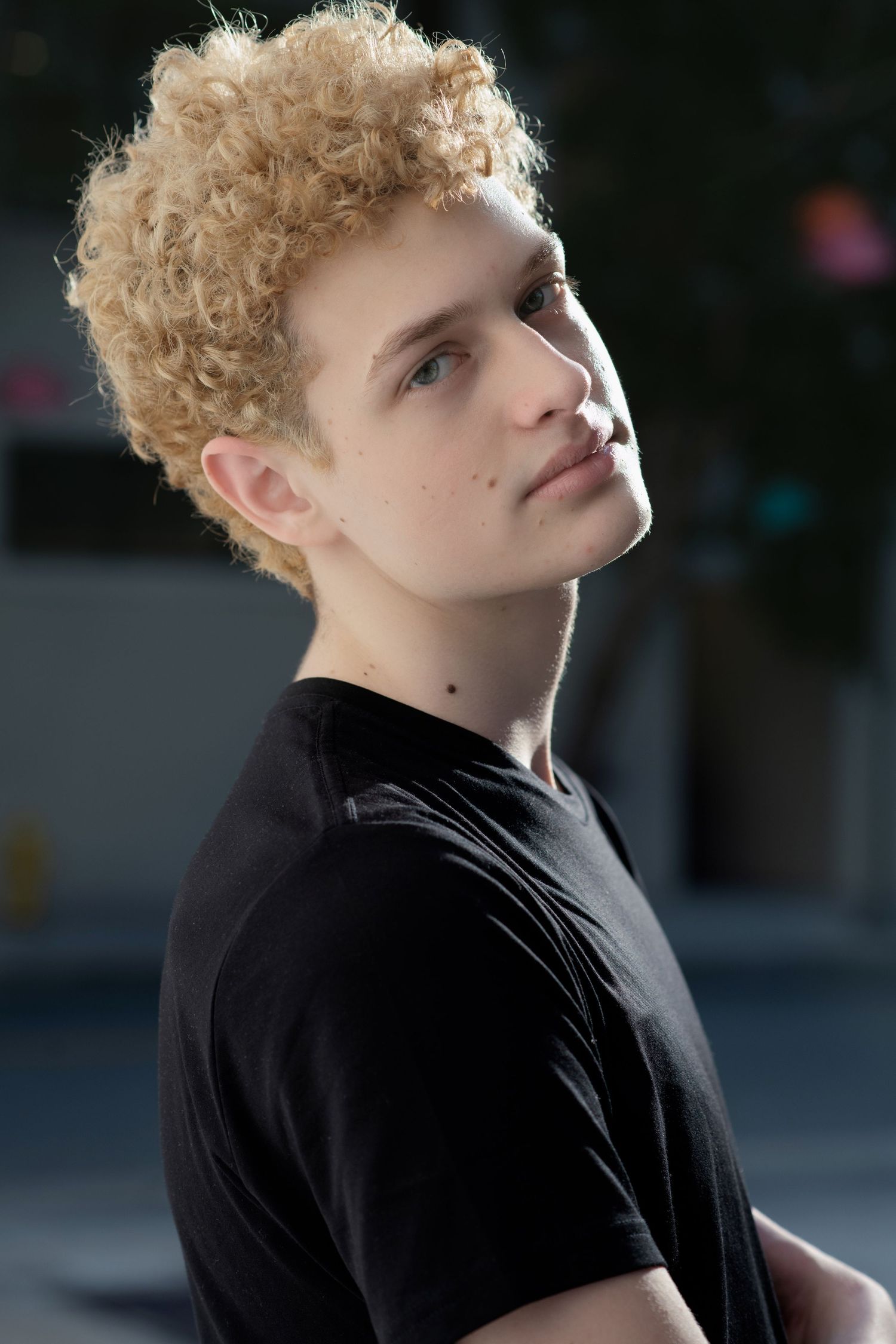 Young man with curly blonde hair wearing black t-shirt poses outdoors in natural sunlight.