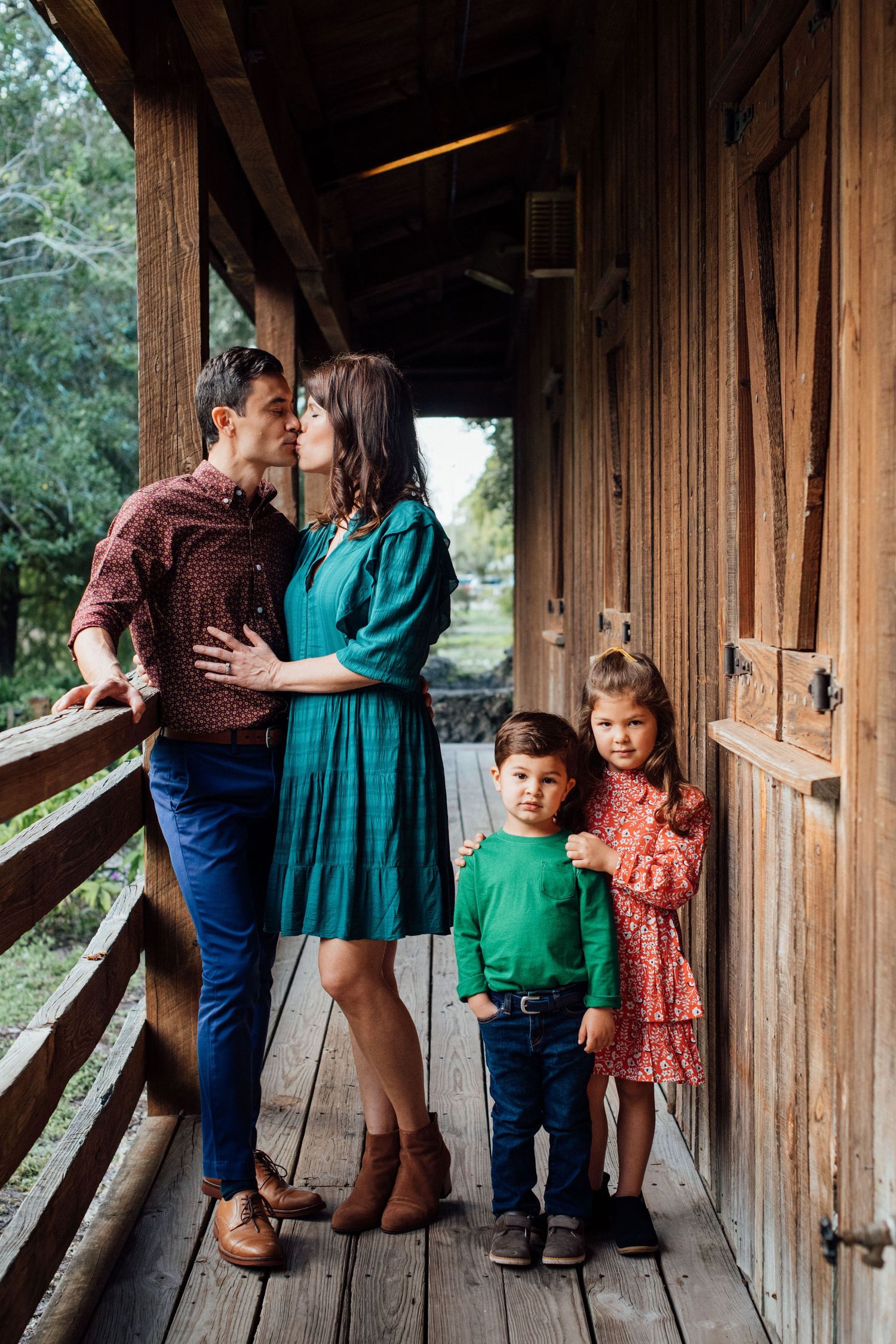 A family of four poses on a wooden porch, parents kissing while children stand beside them.